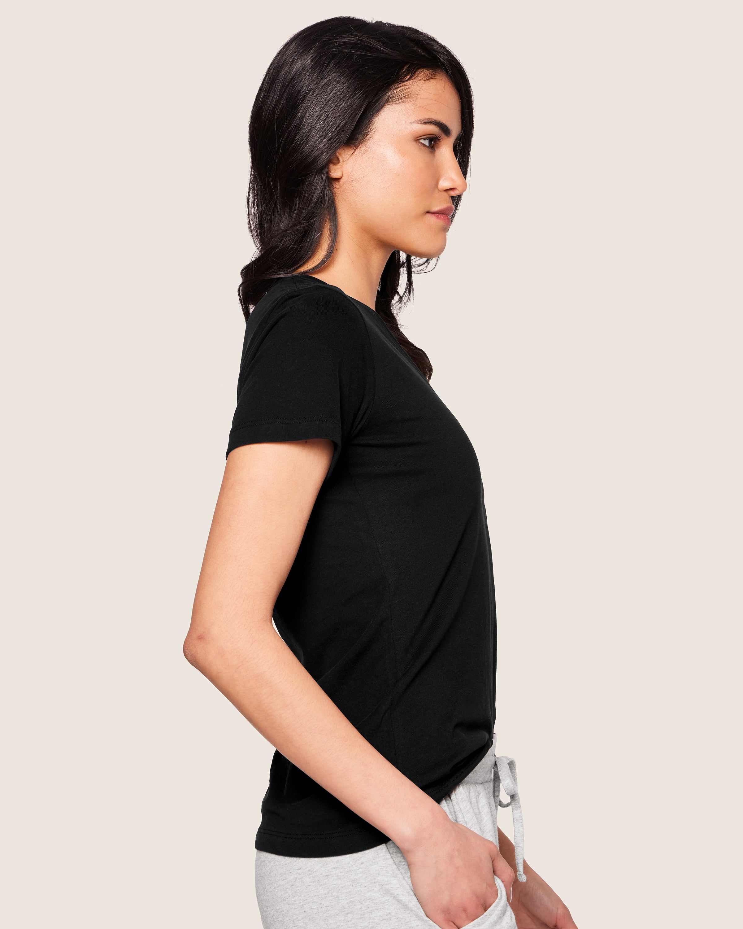 A woman with long dark hair stands in profile wearing a Womens Pima Short Sleeve Top in Black by Petite Plume and light gray sweatpants made from exquisite Peruvian Pima cotton, set against a plain, light background.