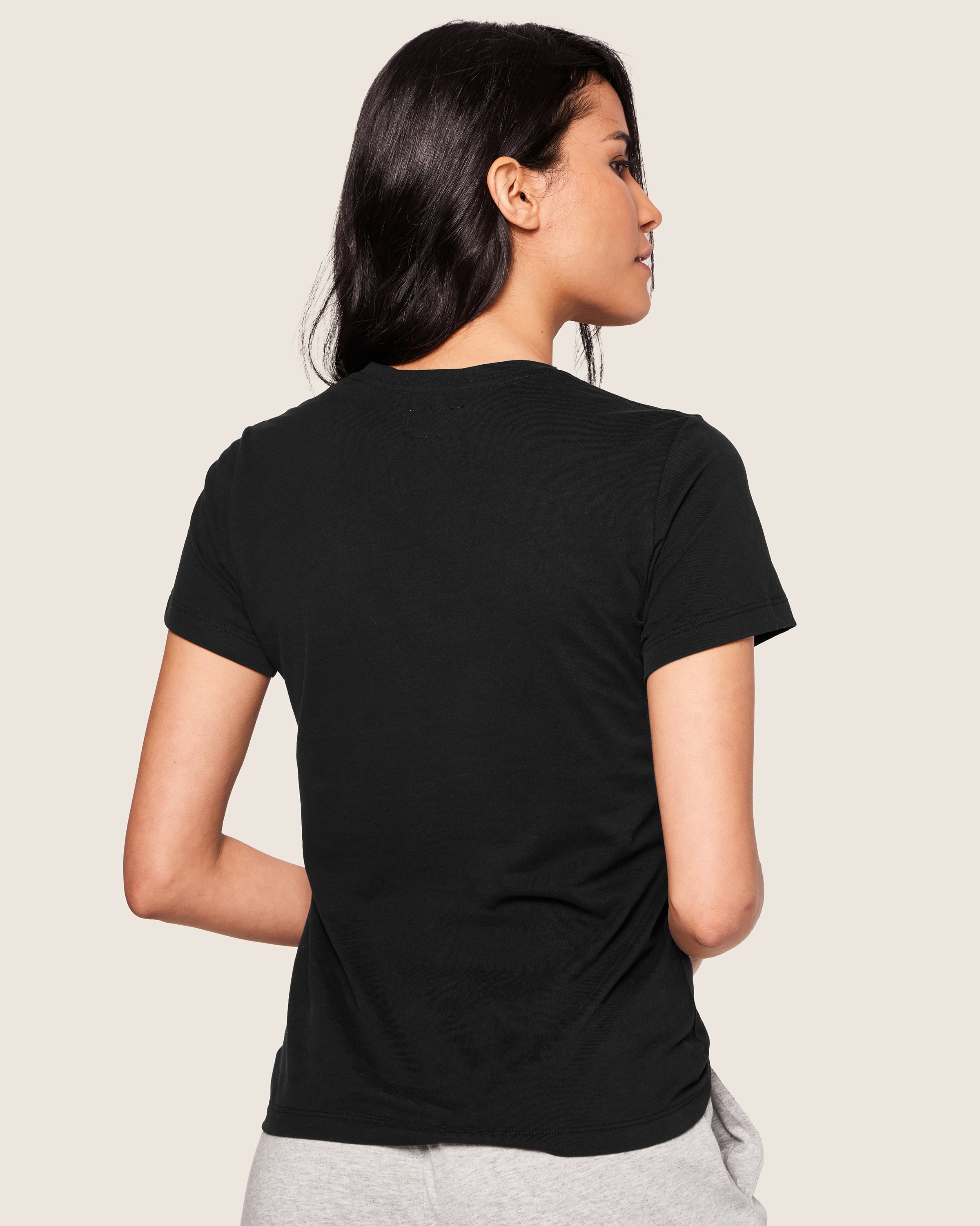 A person with long dark hair is wearing a Womens Pima Short Sleeve Top in Black by Petite Plume and light gray pants, both crafted from soft Peruvian Pima cotton. They stand with their back to the camera against a plain beige background.