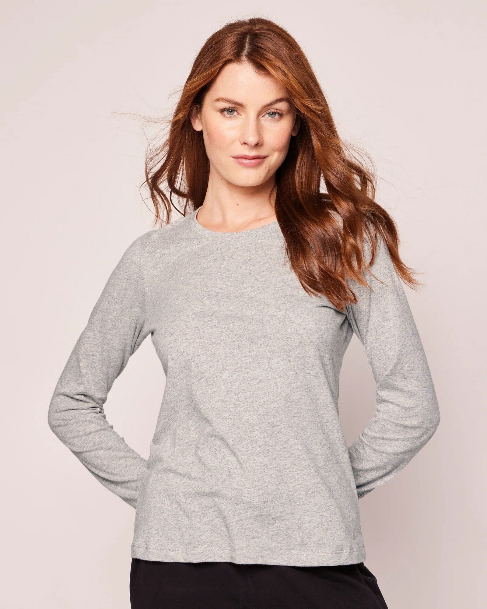 A woman with long, wavy brown hair is wearing a Womens Pima Long Sleeve Top in Light Grey by Petite Plume and black pants. Her hands are tucked into her pockets against a plain background, radiating relaxed elegance reminiscent of luxury pajamas.