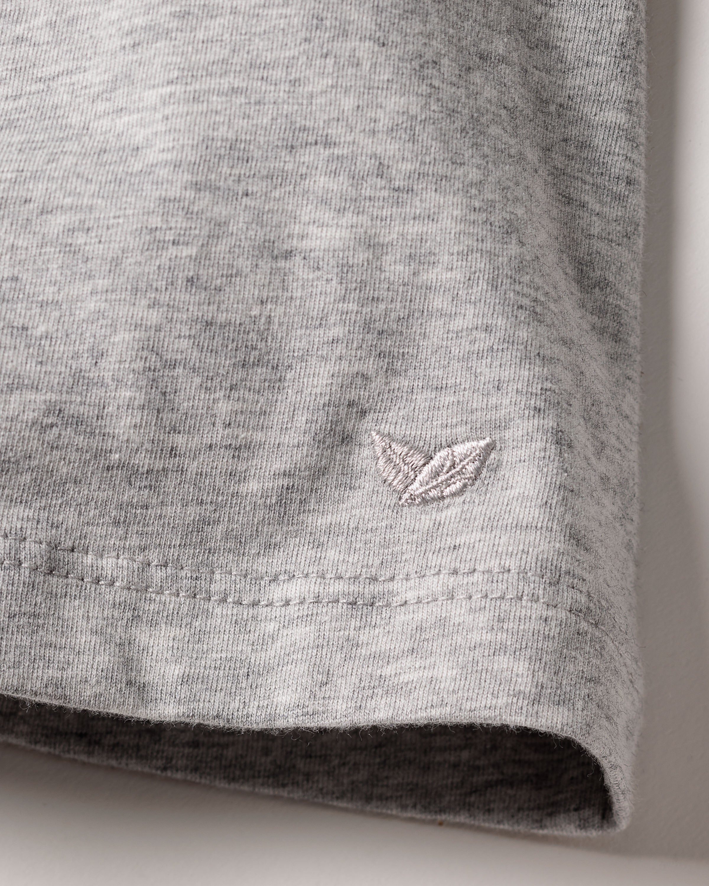Close-up of Petite Plumes Womens Pima Tank Top in Light Grey, featuring a soft Peruvian Pima cotton heathered knit with an embroidered two-leaf emblem. The neatly stitched hem indicates a high-quality garment.