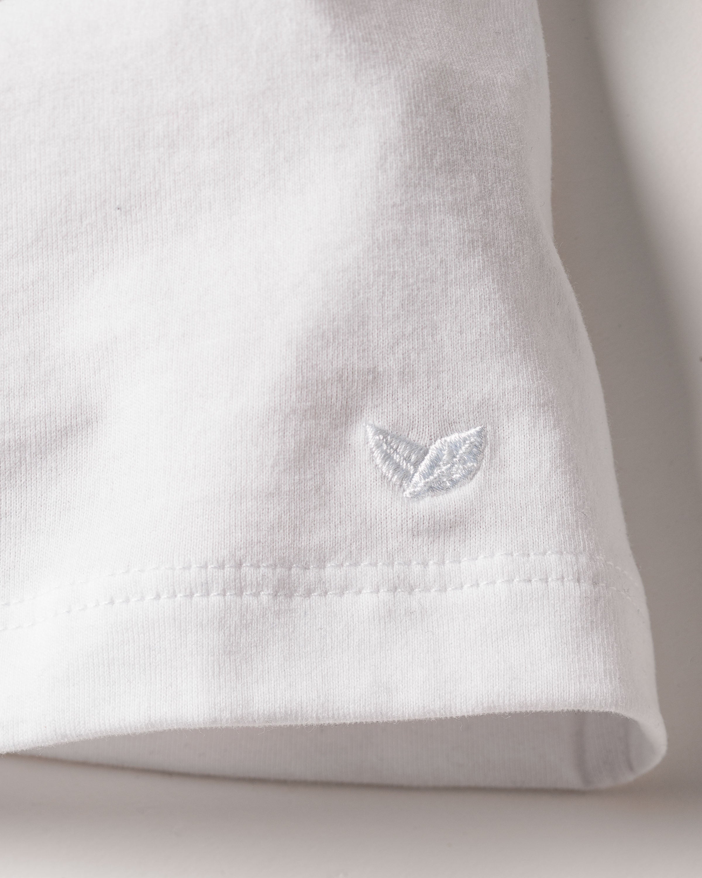 A close-up of a Womens Pima Tank Top in White by Petite Plume showcases luxurious Peruvian Pima cotton with a subtle embroidered leaf logo. The stitching and texture are visible, and soft lighting accentuates its clean, simple design.