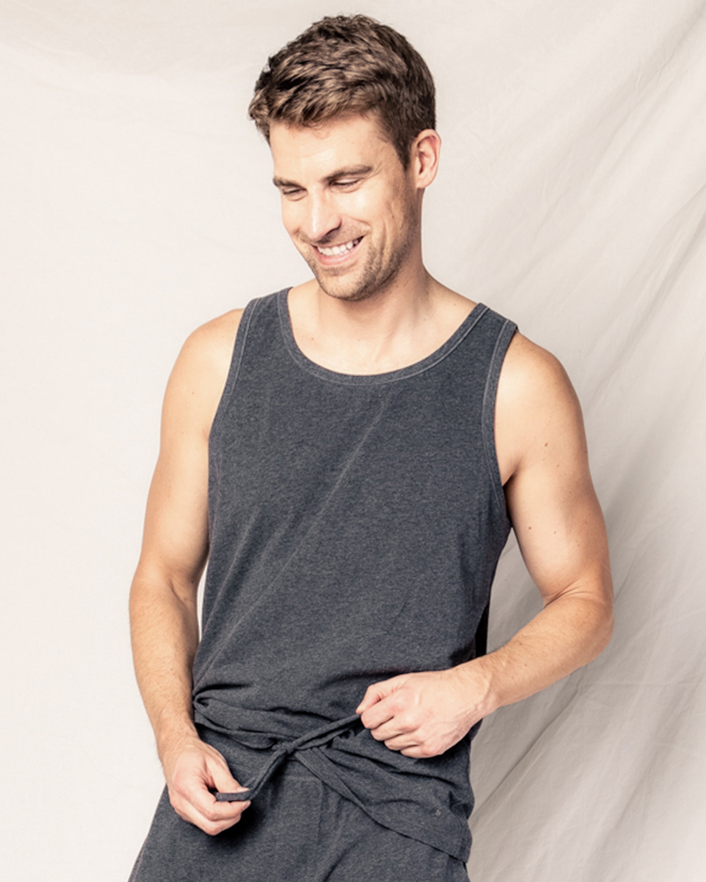 A smiling man with short brown hair, wearing the Petite Plume Mens Pima Tank Top in Dark Heather Grey, stands against a light background, gently holding the shirts hem while looking slightly downwards.
