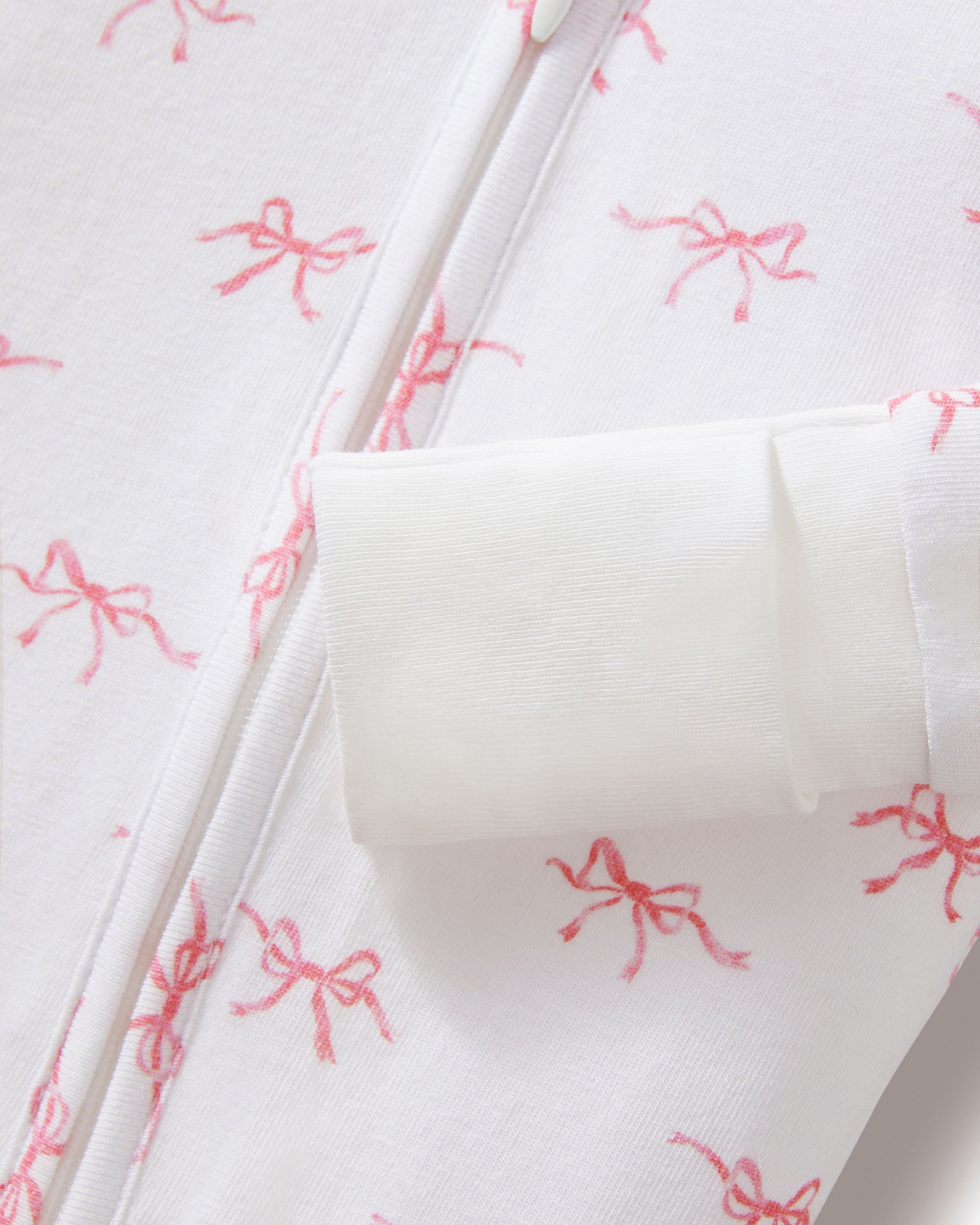 The Baby's Pima Peekaboo Romper in Blushing Bows is a white onesie with long sleeves and legs, accented by white cuffs and an angled zipper, and features a pink bow pattern.
