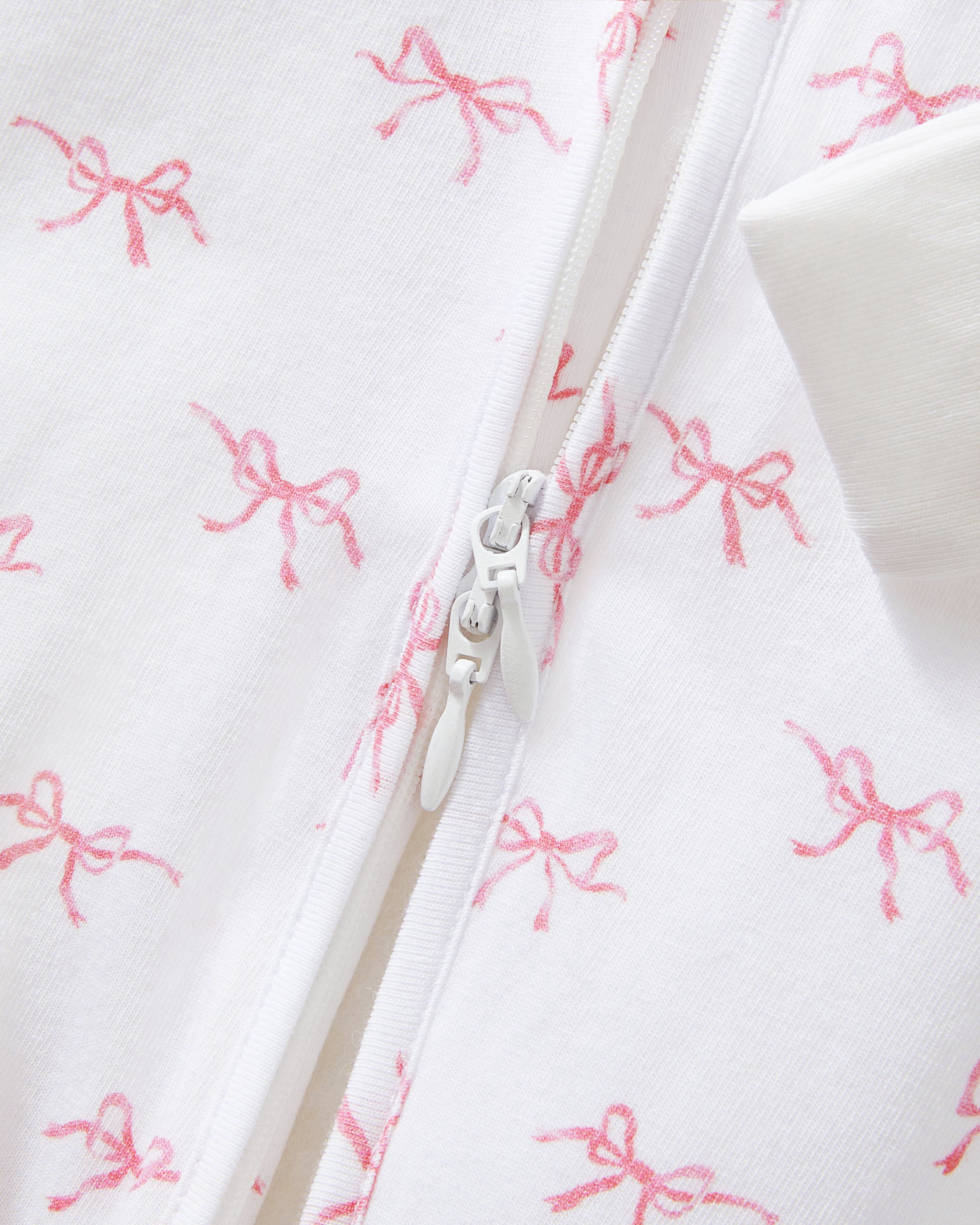 The Baby's Pima Peekaboo Romper in Blushing Bows is a white onesie with long sleeves and legs, accented by white cuffs and an angled zipper, and features a pink bow pattern.