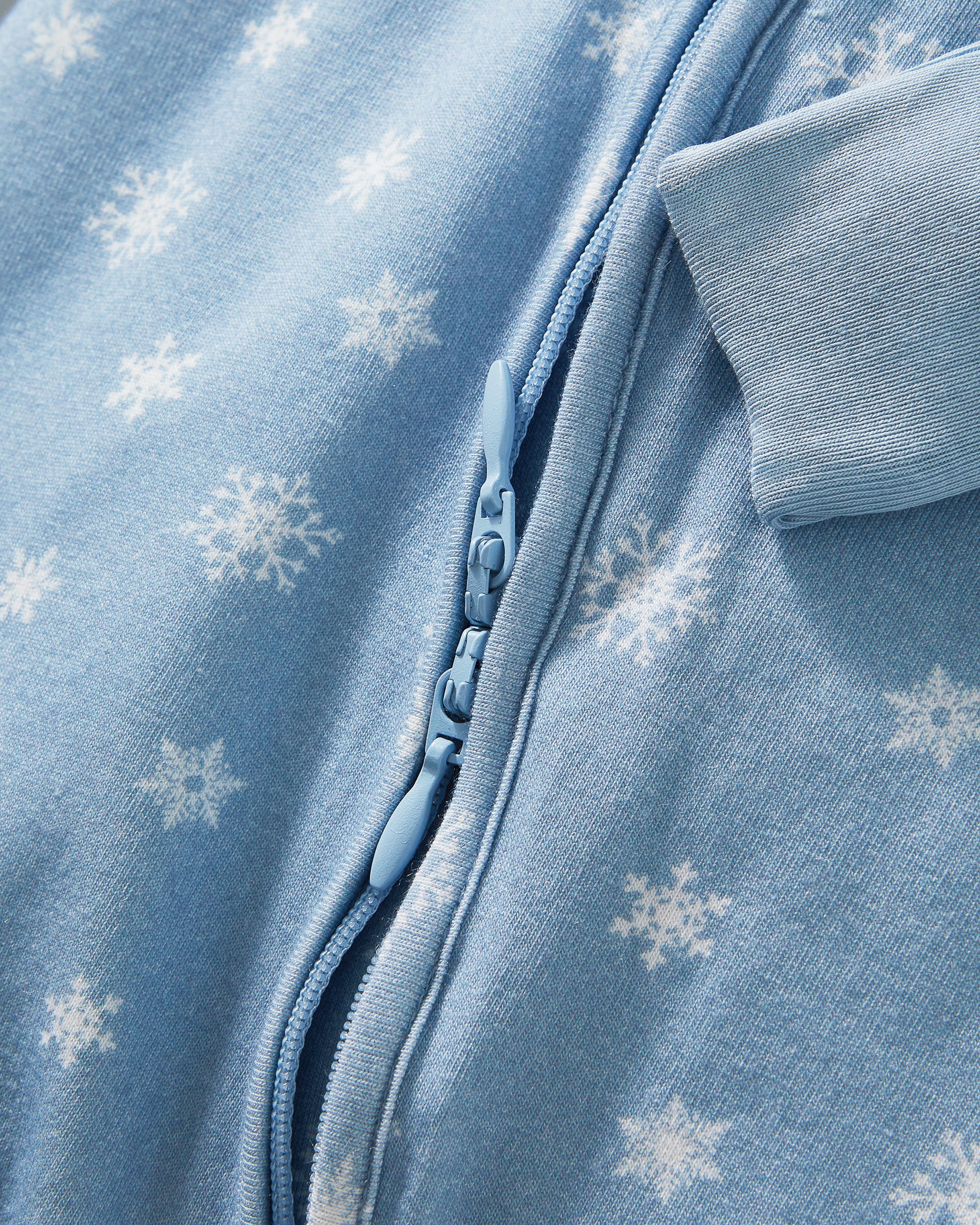 The Baby's Pima Peekaboo Romper in Periwinkle Wonderland features a light blue snowflake pattern, long sleeves with fold-over cuffs, and a diagonal zipper from neckline to left ankle; one sleeve and one leg are folded.
