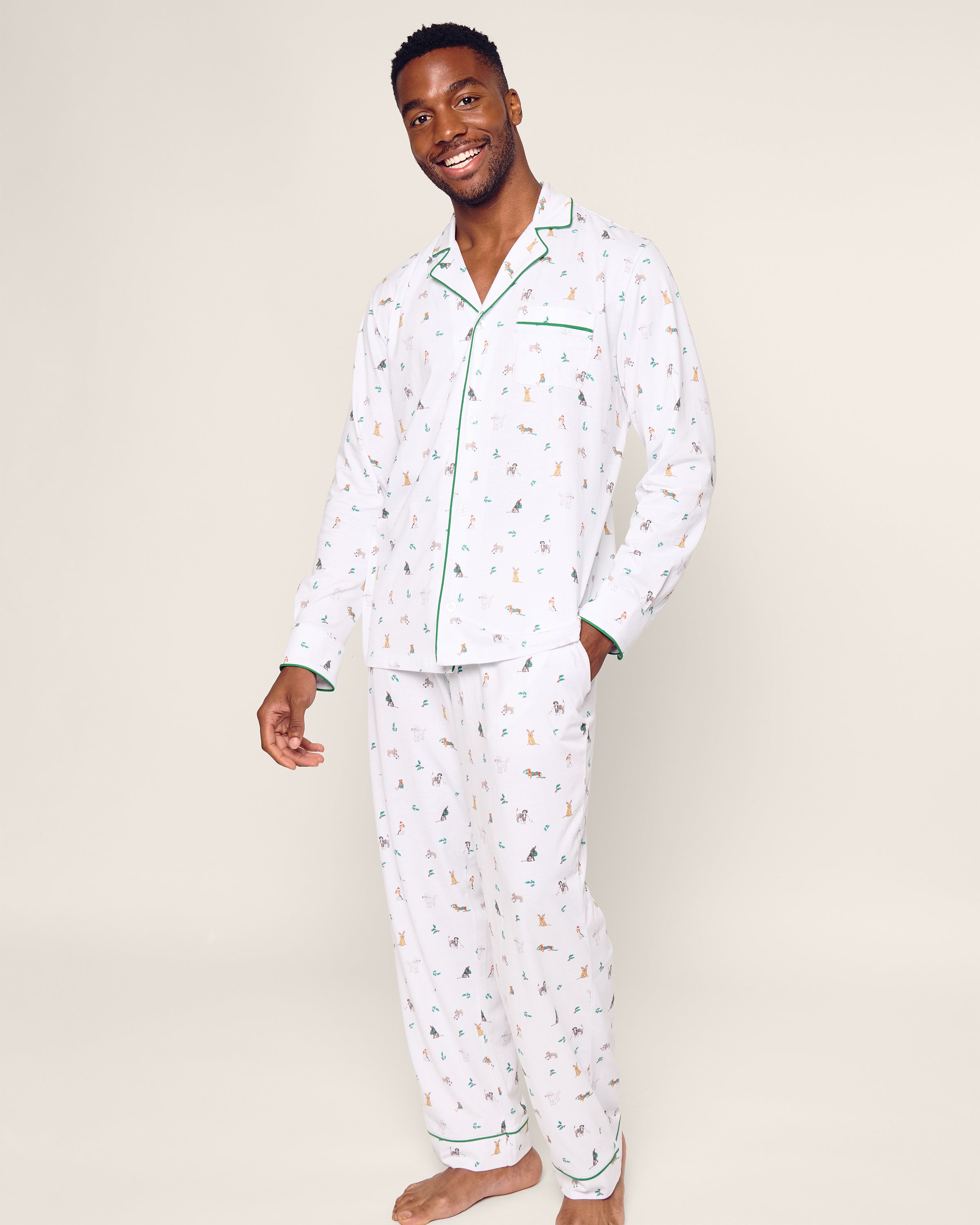 A person smiles in Petite Plumes Mens Pima Pajama Set in Jingle Paws, featuring colorful animal designs on soft Peruvian Pima cotton with green piping and a chest pocket against a plain, light background, enhancing their cheerful appearance.