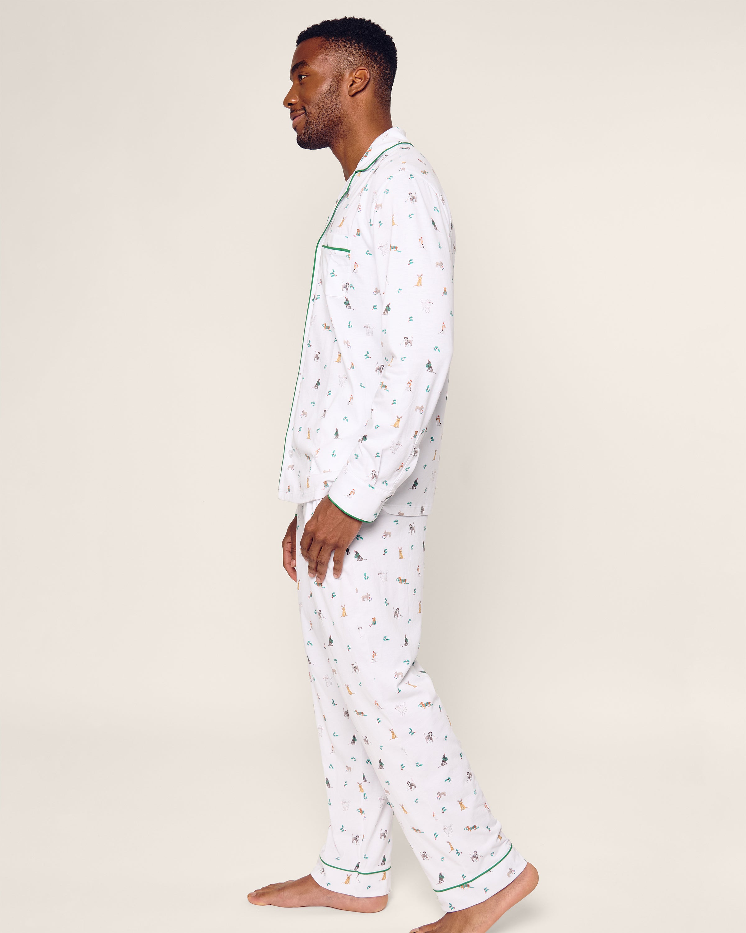 Standing barefoot, a person wears the luxurious Mens Pima Pajama Set in Jingle Paws by Petite Plume, crafted from Peruvian Pima cotton. The white pajamas with colorful patterns feature green piping and a button-up top against a simple light background, perfect for holiday sleepwear.