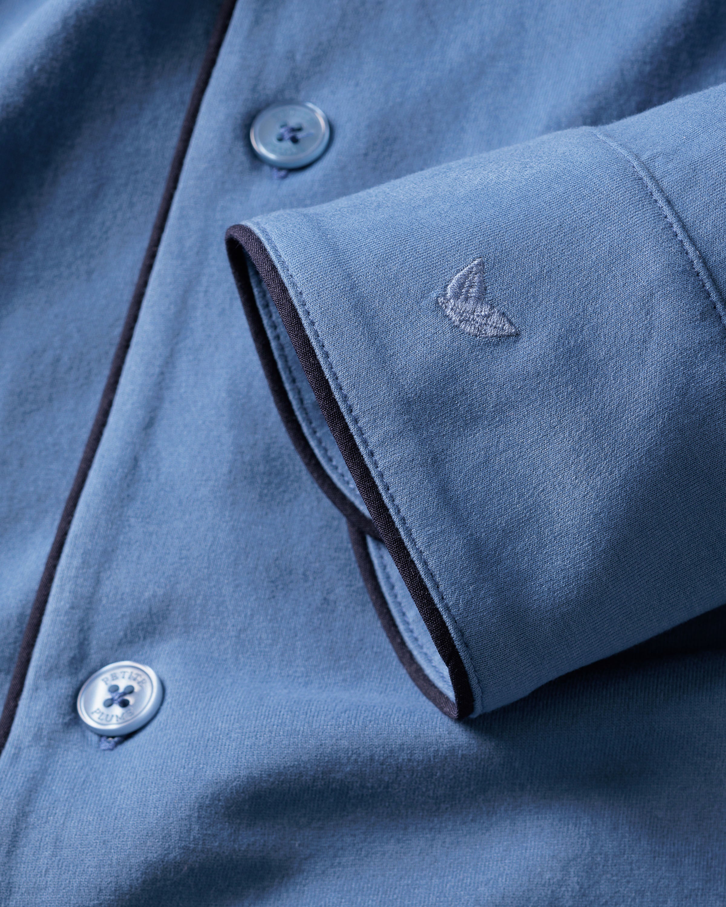 Close-up of Petite Plumes Mens Pima Pajama Set in Mystique Blue features a folded sleeve with an embroidered logo, subtly patterned buttons, and dark fabric piping, embodying luxury sleepwear.