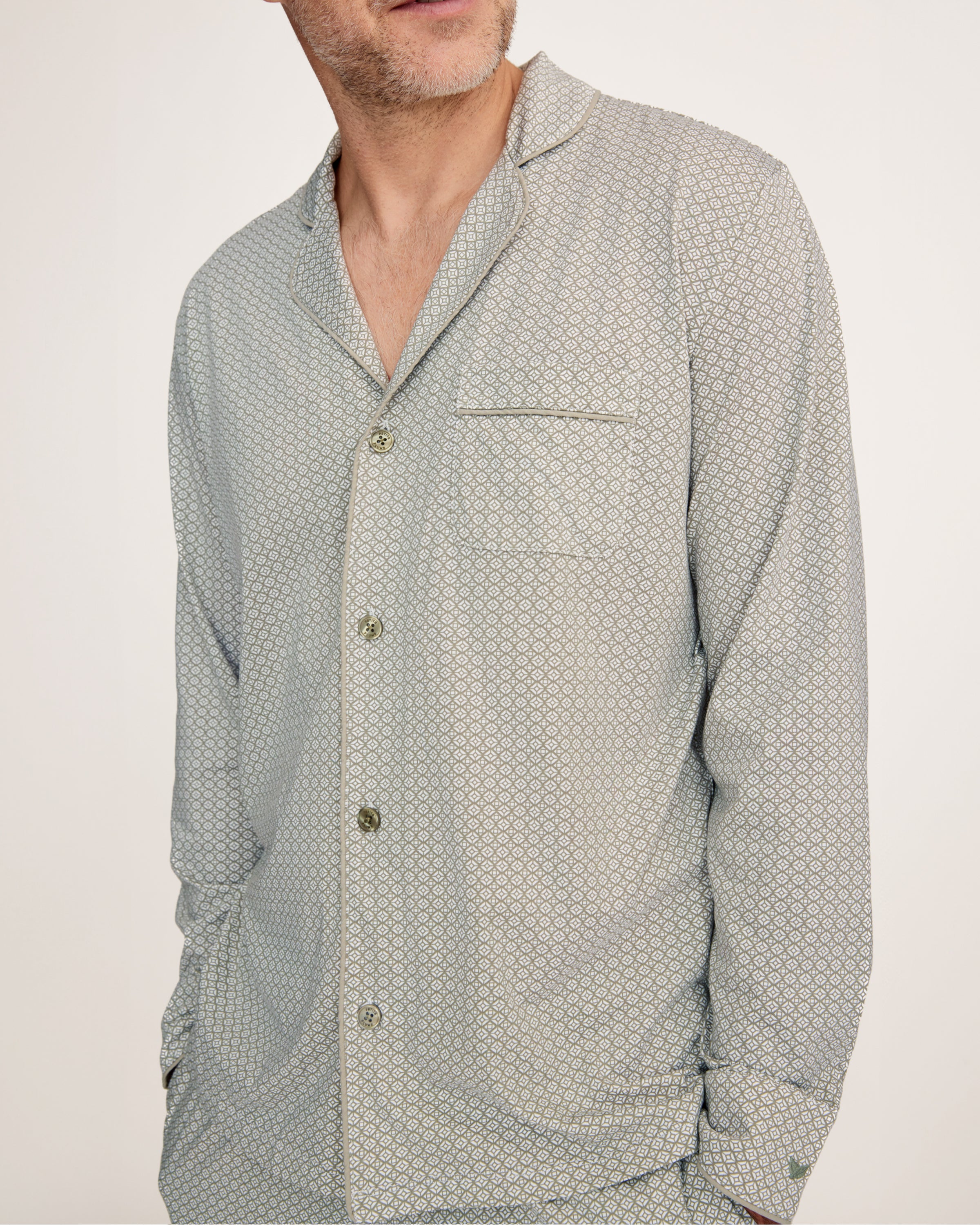 A man with short gray hair and light stubble stands barefoot in the Men's Lightweight Pima Pajama Set in Sage Geo against a white background, gazing to the side with a relaxed expression.