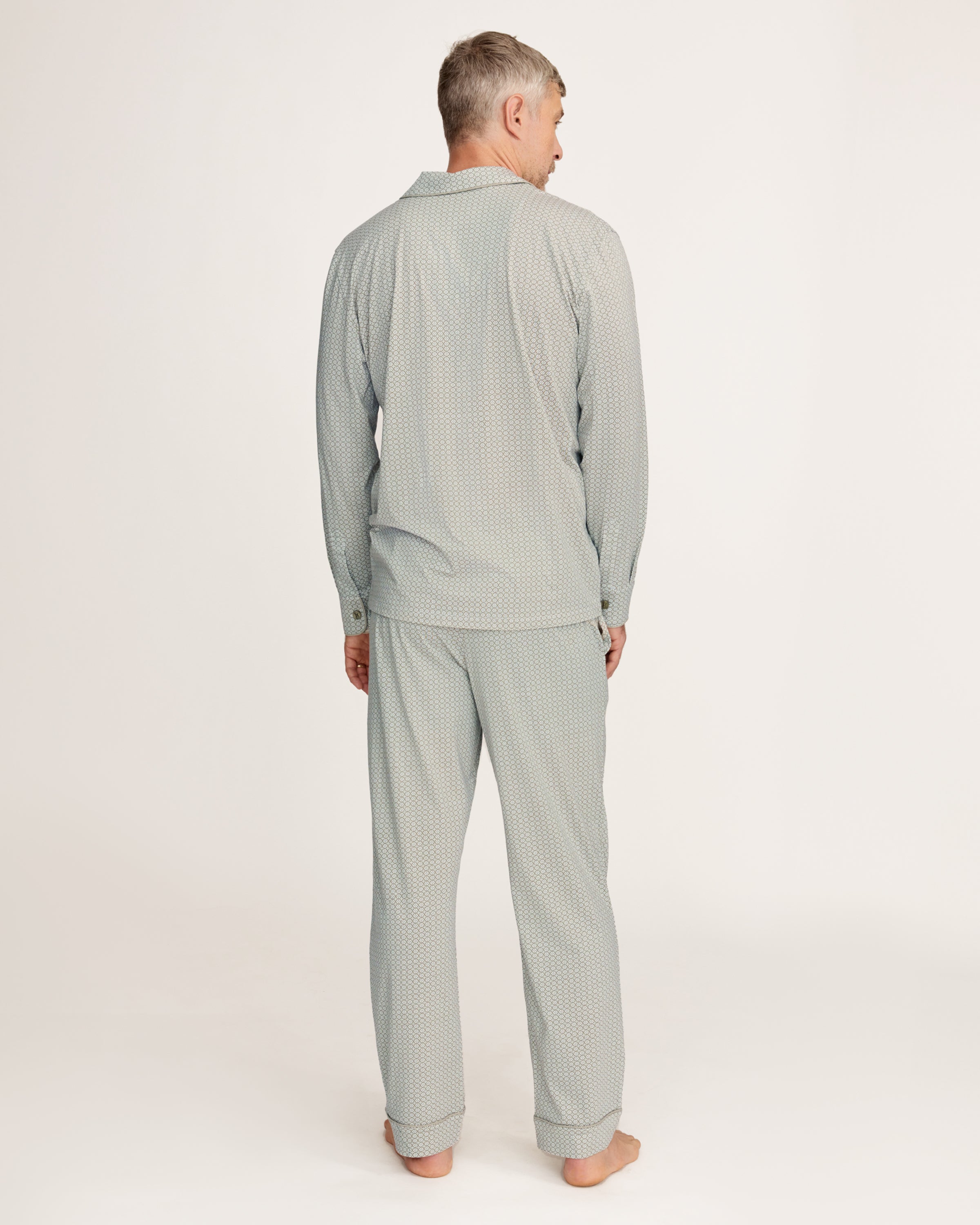 A man with short gray hair and light stubble stands barefoot in the Men's Lightweight Pima Pajama Set in Sage Geo against a white background, gazing to the side with a relaxed expression.