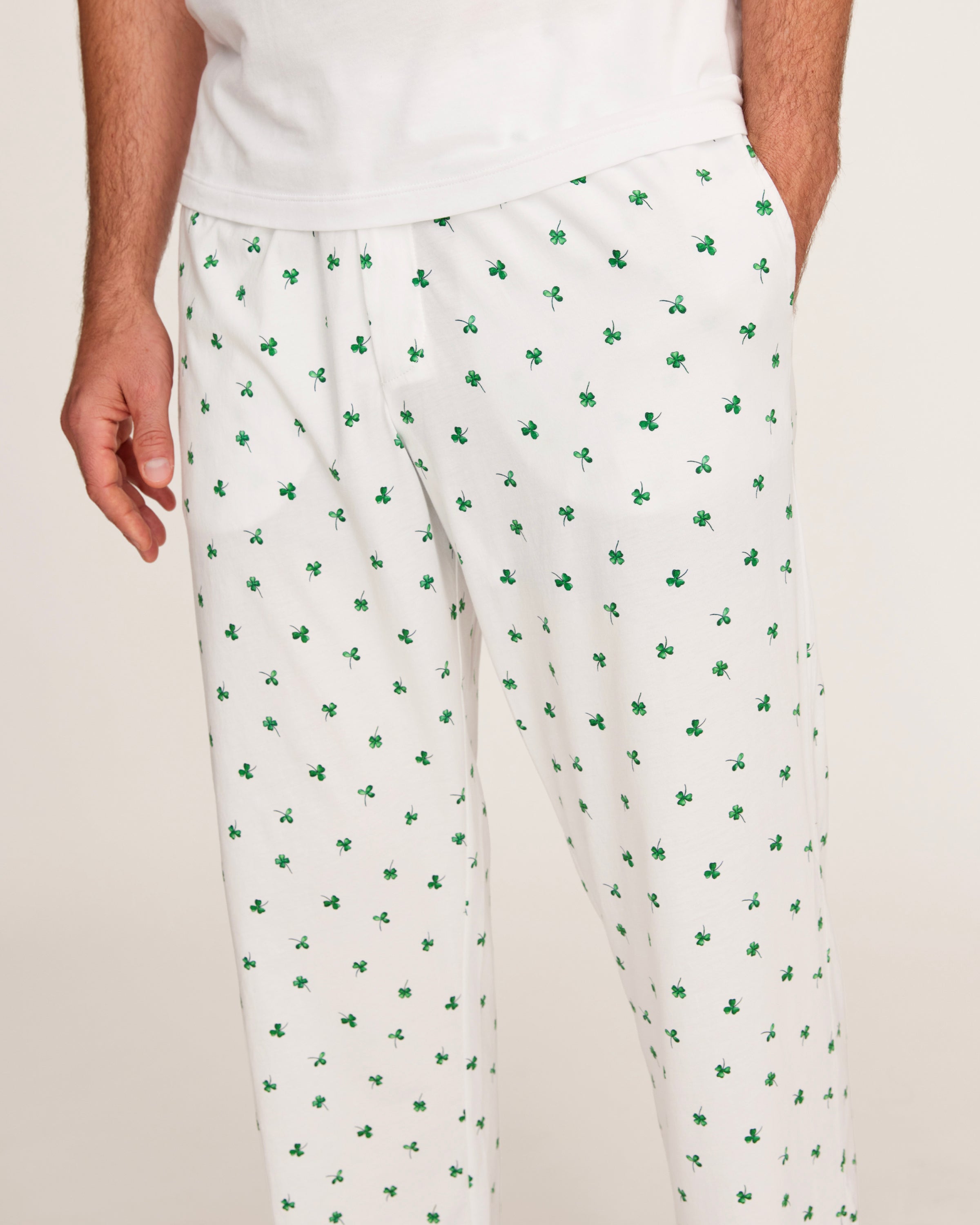 A man with short gray hair stands barefoot against a plain light background, wearing a plain white tee and Men's Pima Pants in Shamrocks—a white pajama pant with a small green pattern.