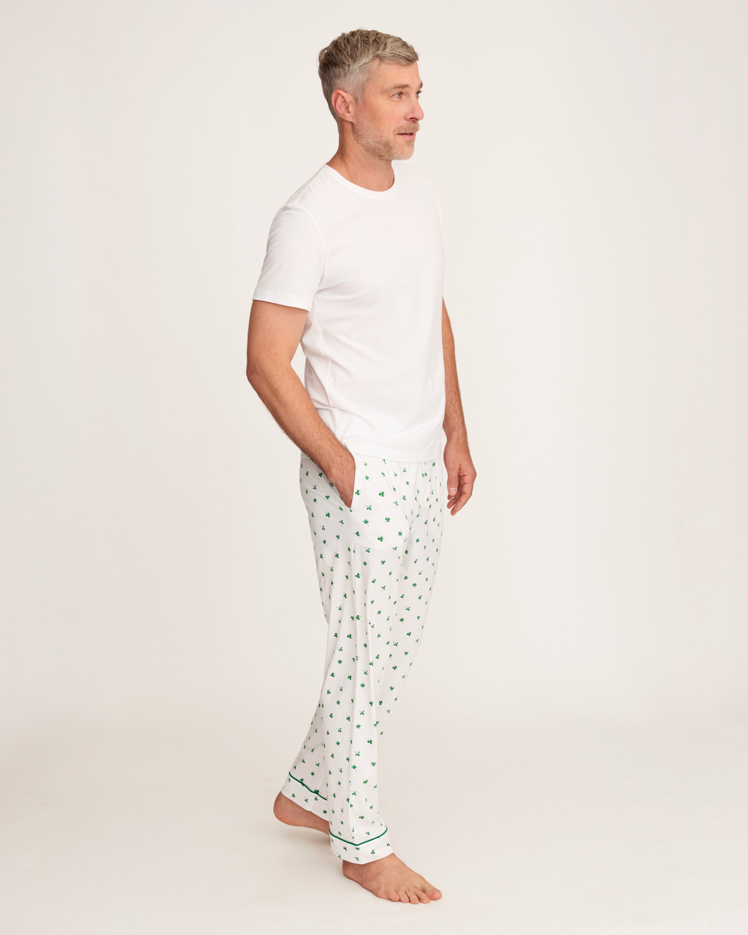 A man with short gray hair stands barefoot against a plain light background, wearing a plain white tee and Men's Pima Pants in Shamrocks—a white pajama pant with a small green pattern.