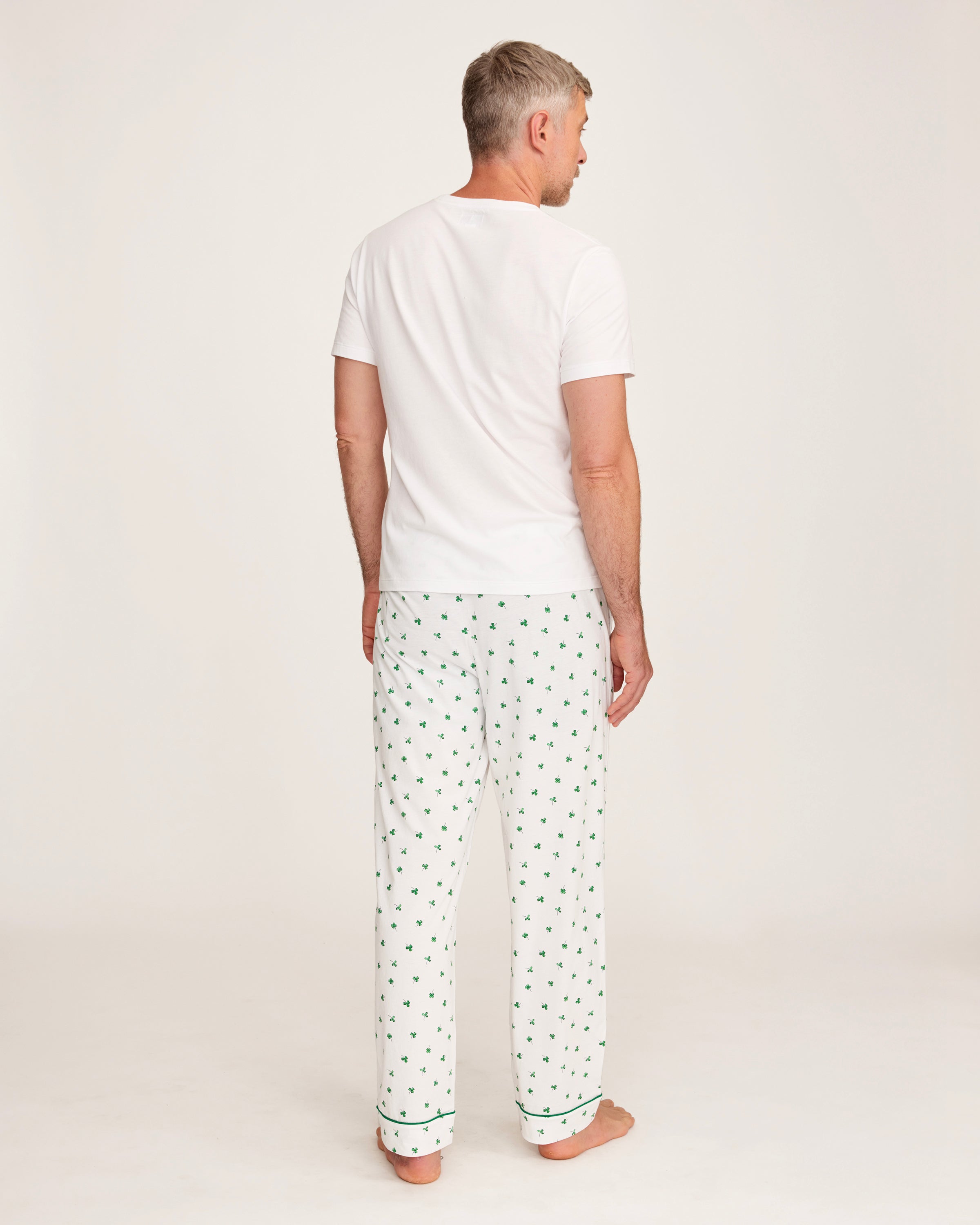 A man with short gray hair stands barefoot against a plain light background, wearing a plain white tee and Men's Pima Pants in Shamrocks—a white pajama pant with a small green pattern.