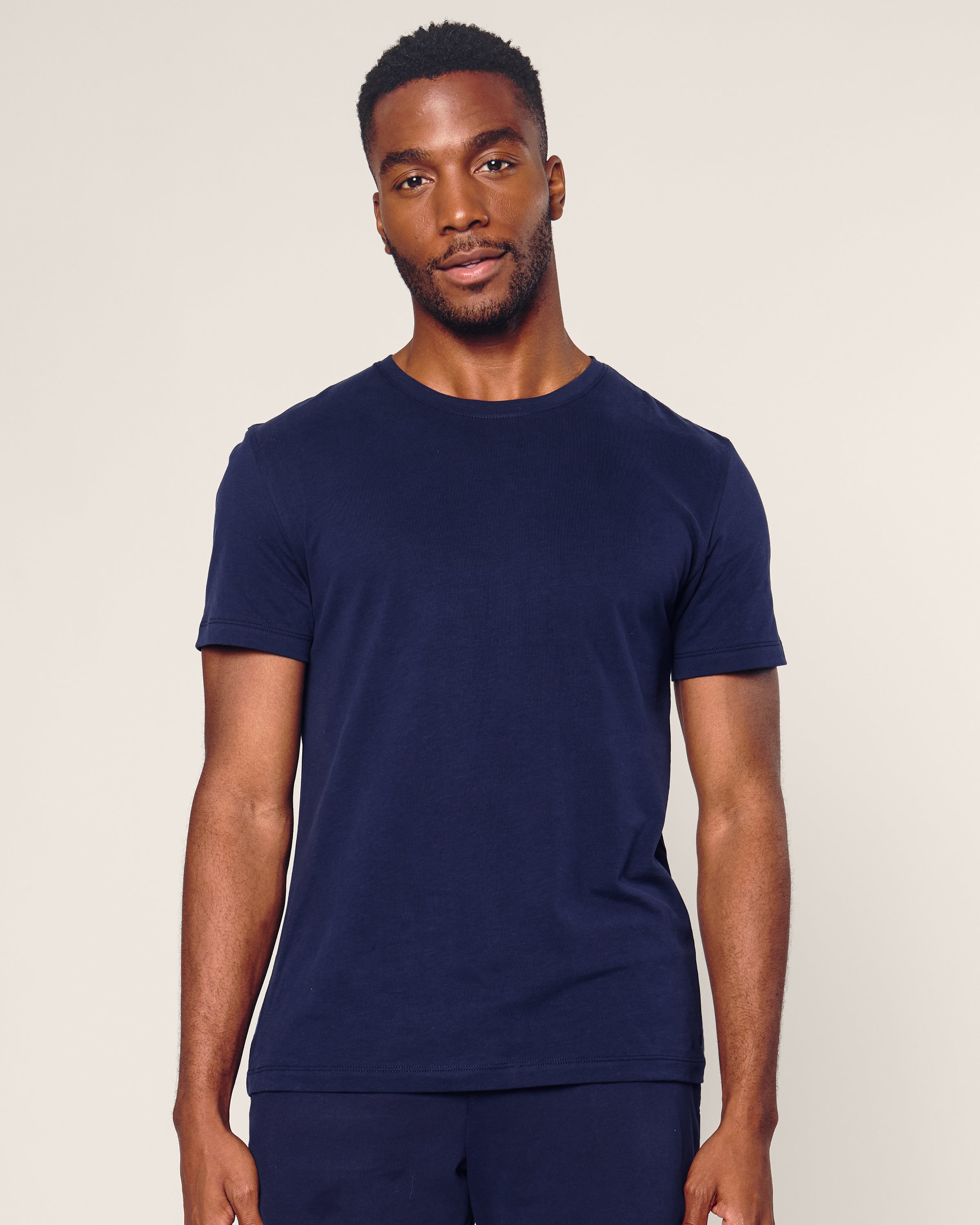 A person wearing Petite Plumes Mens Pima Crew Neck Short Sleeve Top in Navy, reminiscent of soft Peruvian Pima cotton, stands against a light gray background. They have short hair and facial hair with a calm, relaxed expression.