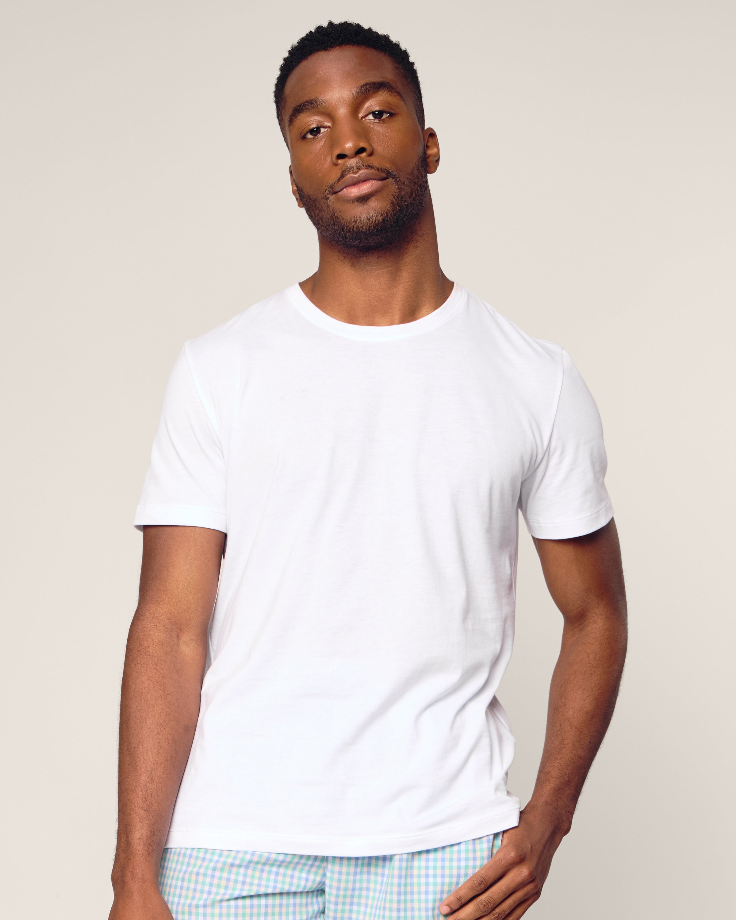 A man wearing a plain white Mens Pima Crew Neck Short Sleeve Top by Petite Plume and light-colored pants stands against a neutral background, looking at the camera with a relaxed expression. His right hand rests gently on his hip, exuding comfort akin to jersey knit pajamas.