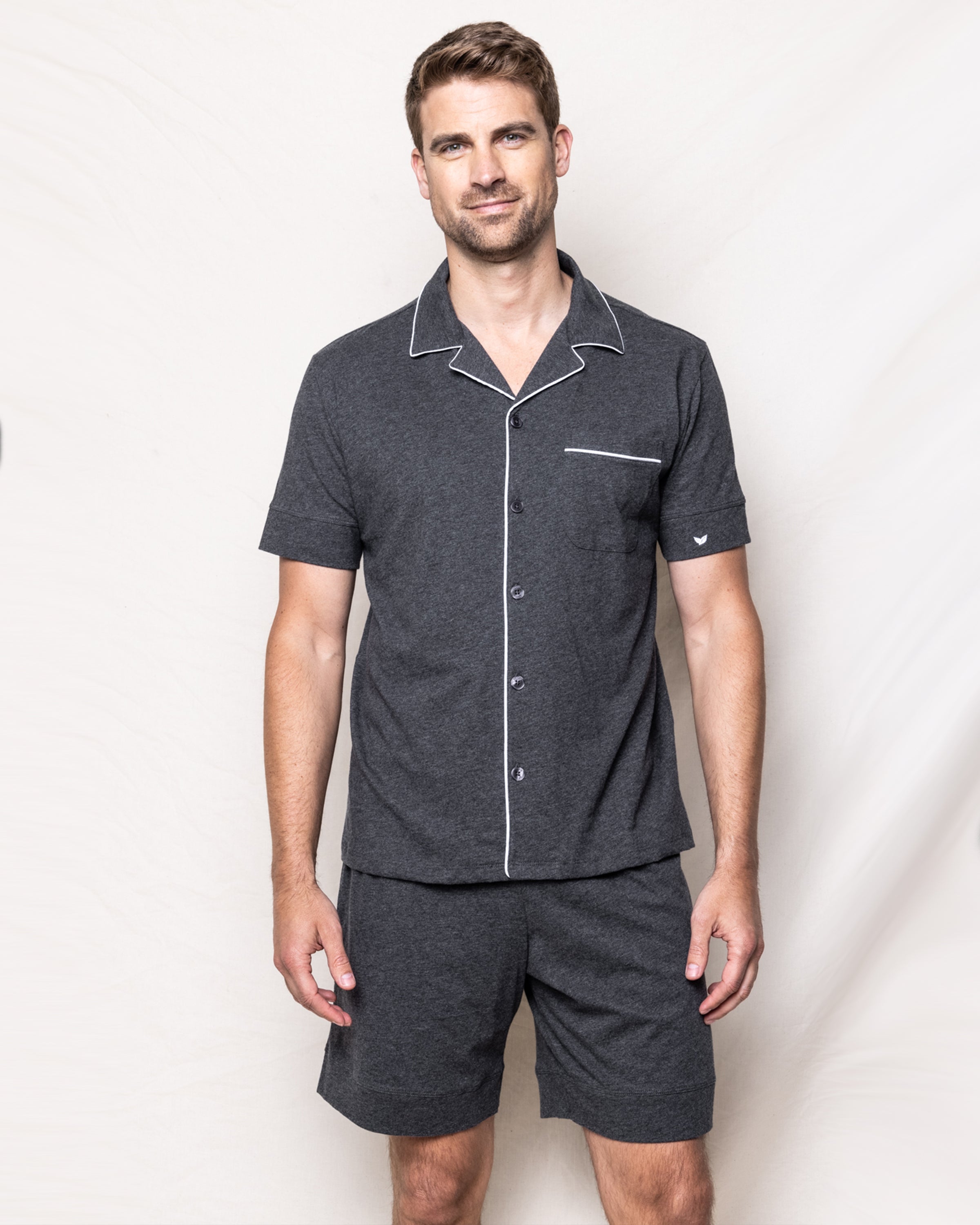 A man is wearing the Mens Pima Pajama Short Set in Dark Heather Grey by Petite Plume. Its crafted from yarn-dyed fabric, has white piping detail, a buttoned front, and a chest pocket. Hes standing against a light background with a neutral expression while looking at the camera.