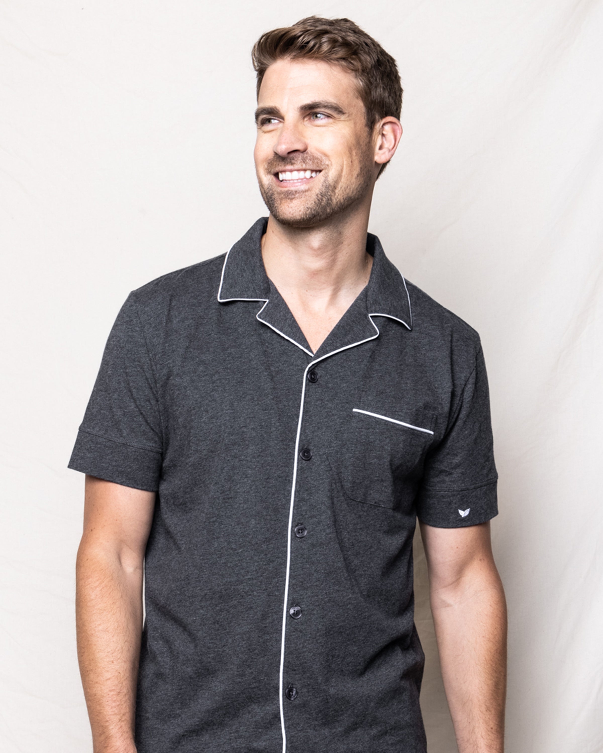A man with short brown hair and light facial hair wears the Mens Pima Pajama Short Set in Dark Heather Grey by Petite Plume. He is smiling and looking to the side against a plain, light background.