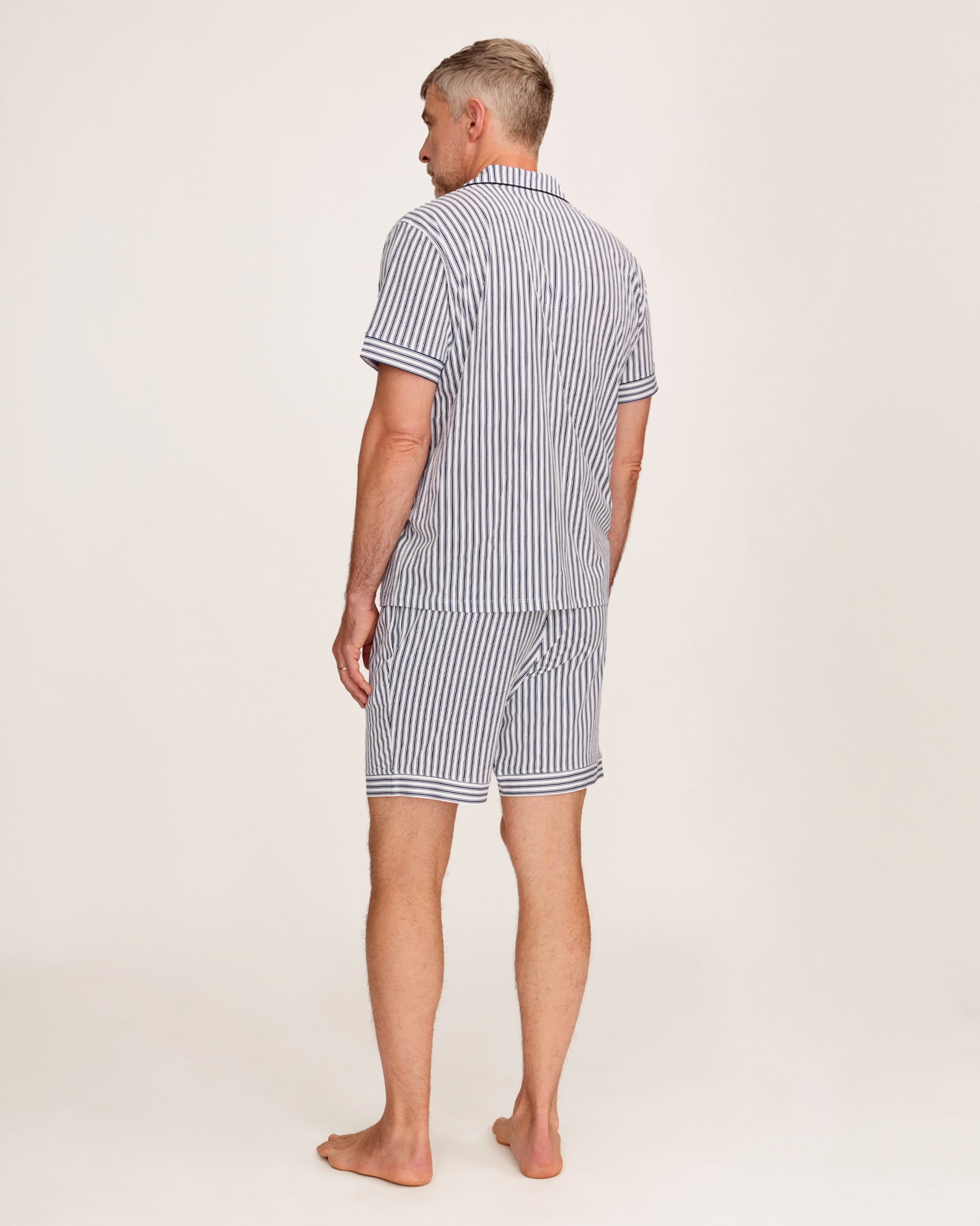 A man stands barefoot in a studio wearing the Men's Pima Pajama Short Set in Navy French Ticking, which features vertical blue and white stripes. He looks slightly to the side with one hand in his pocket.