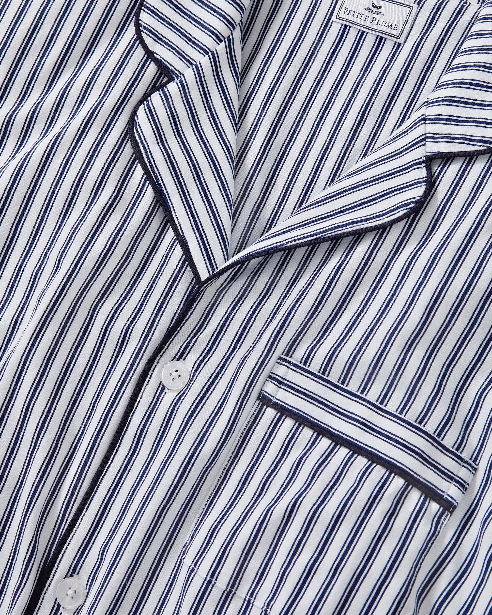 A man stands barefoot in a studio wearing the Men's Pima Pajama Short Set in Navy French Ticking, which features vertical blue and white stripes. He looks slightly to the side with one hand in his pocket.
