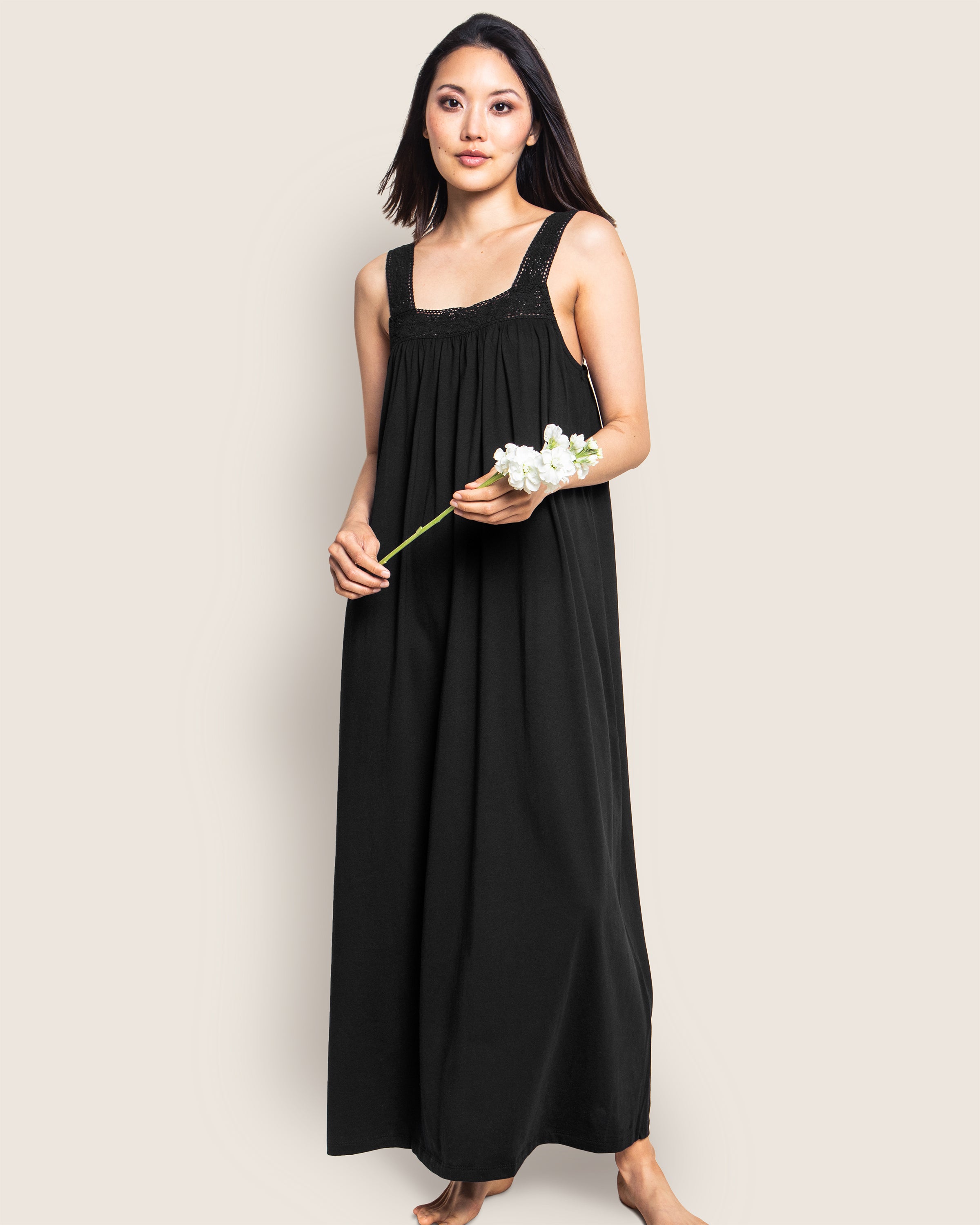 A woman in a Petite Plume Womens Pima Camille Nightgown in Black, stands barefoot holding a white flower, her long dark hair complemented by broad straps and its flowing design as she poses against a plain light background.