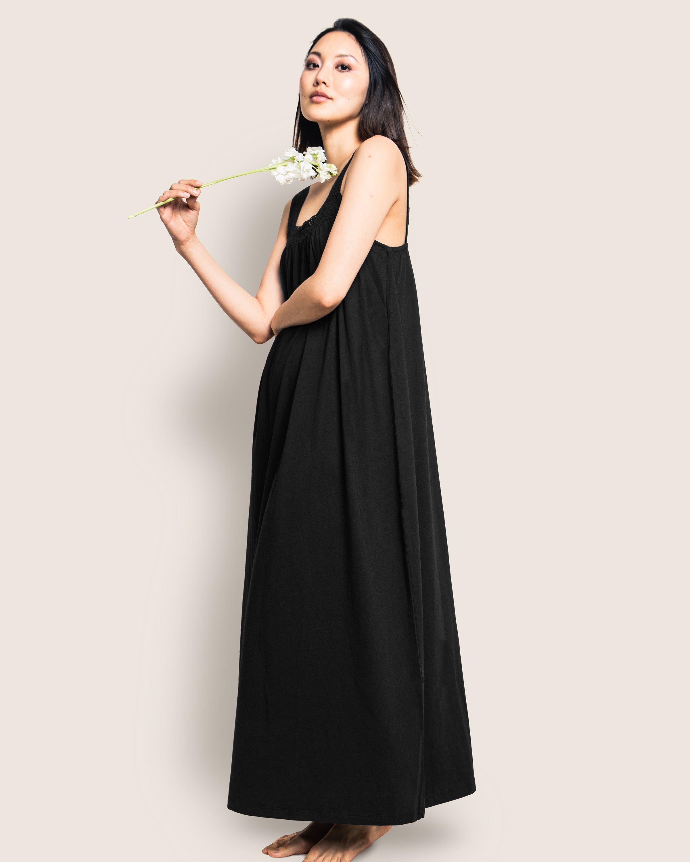 Against a neutral background, a person stands barefoot in a Womens Pima Camille Nightgown in Black by Petite Plume. Holding a white flower, they gaze thoughtfully to the side, their long dark hair cascading down.