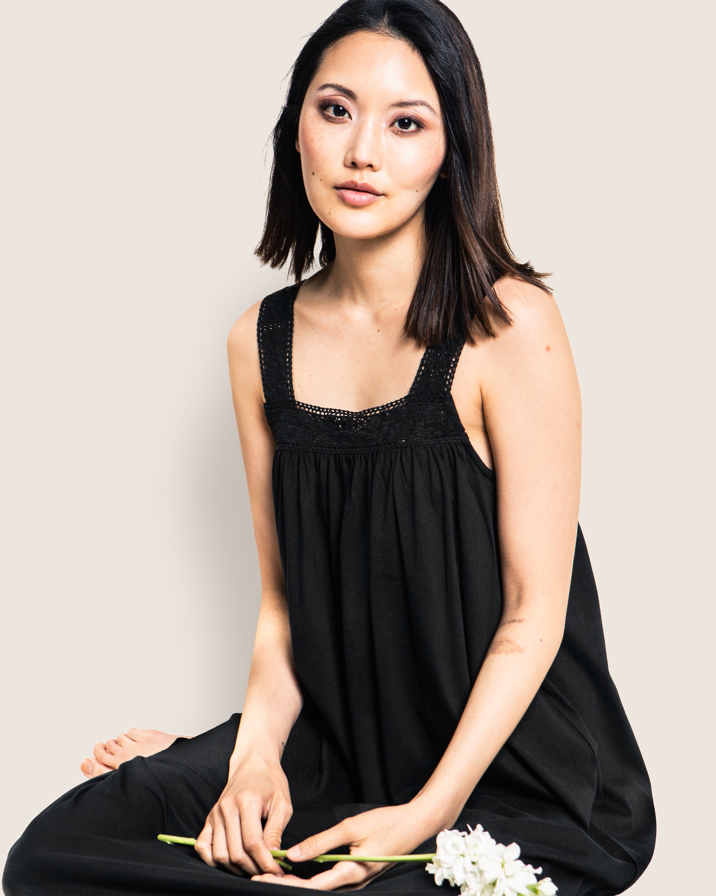 A woman with shoulder-length brown hair sits against a white background, wearing Petite Plumes Womens Pima Camille Nightgown in Black. She gently holds white flowers in one hand, resting them on her knee, while her expression remains calm and composed.