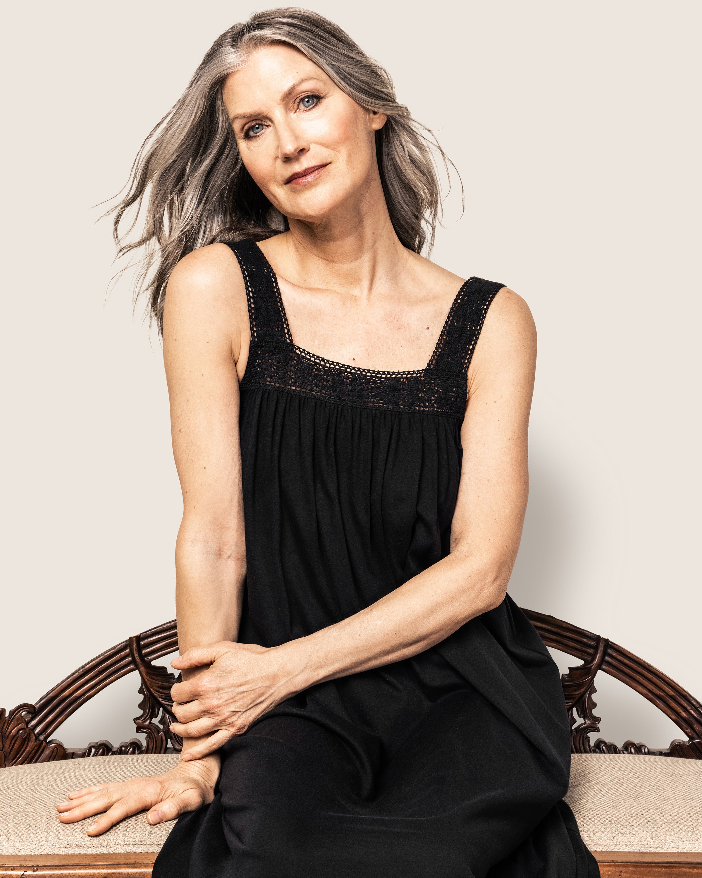 A woman with long gray hair sits serenely on an ornate wooden bench, wearing a Black Womens Pima Camille Nightgown by Petite Plume. Her hands rest gently on her lap against a neutral background.