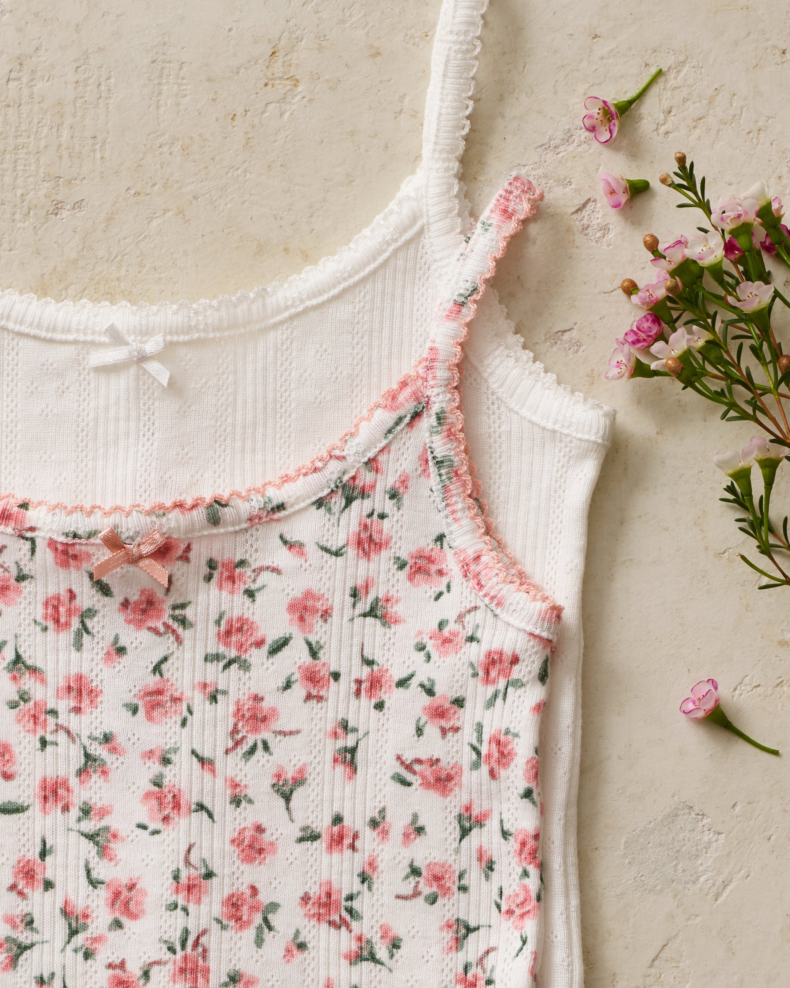 The Kid's Pointelle Cami Set of 2 in White & Pink Floral includes two sleeveless camisoles: one plain white with a bow and one white with a pink and green floral pattern. Both feature thin straps and lace trim.