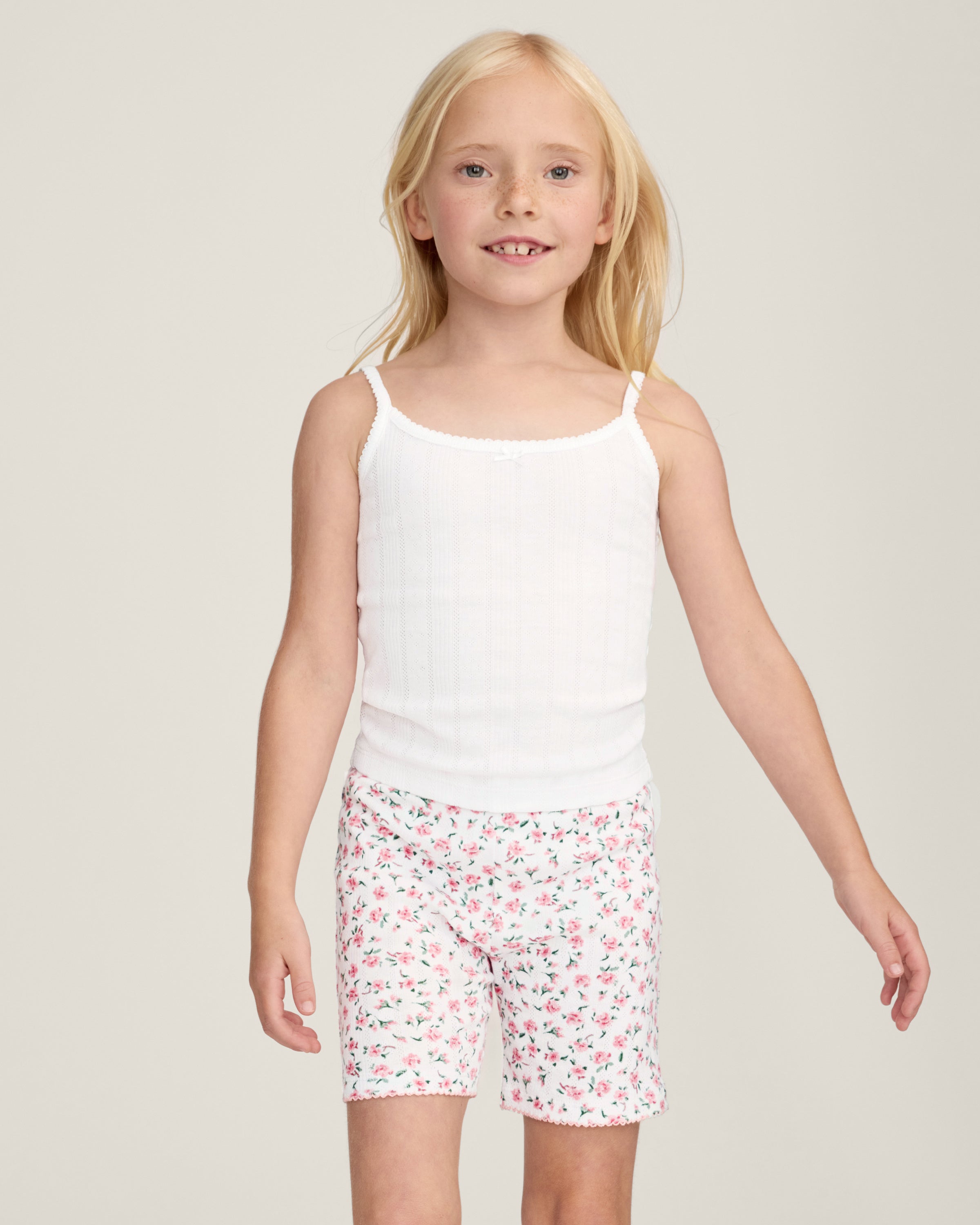 The Kid's Pointelle Cami Set of 2 in White & Pink Floral includes two sleeveless camisoles: one plain white with a bow and one white with a pink and green floral pattern. Both feature thin straps and lace trim.