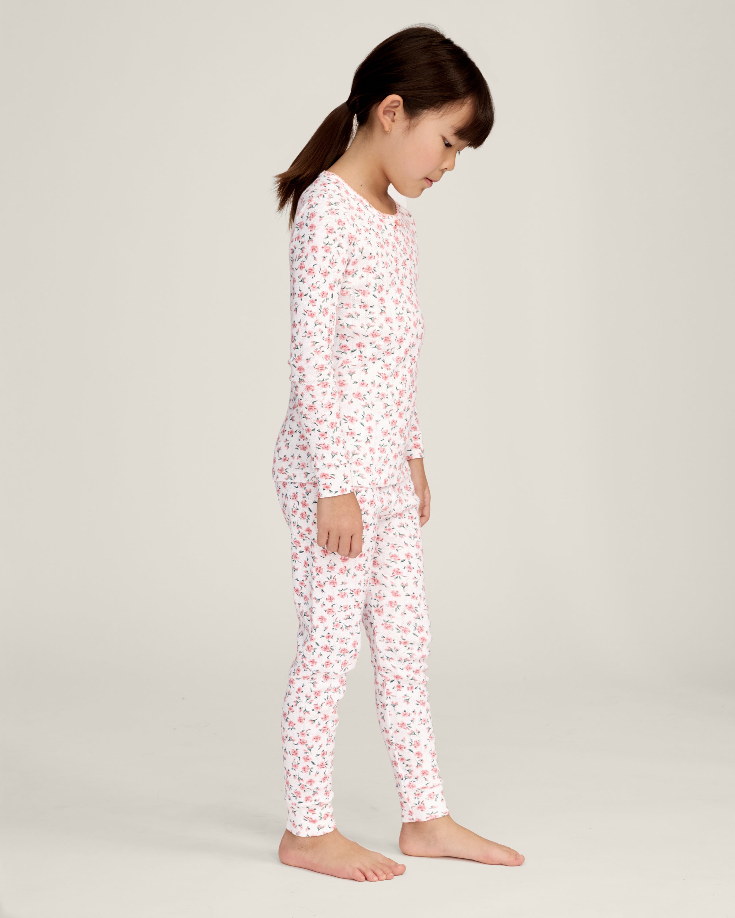 A young girl stands barefoot against a plain background, wearing the Kid's Pointelle Snug Fit Pajama Set in Pink Floral. She faces forward with a relaxed, neutral expression.