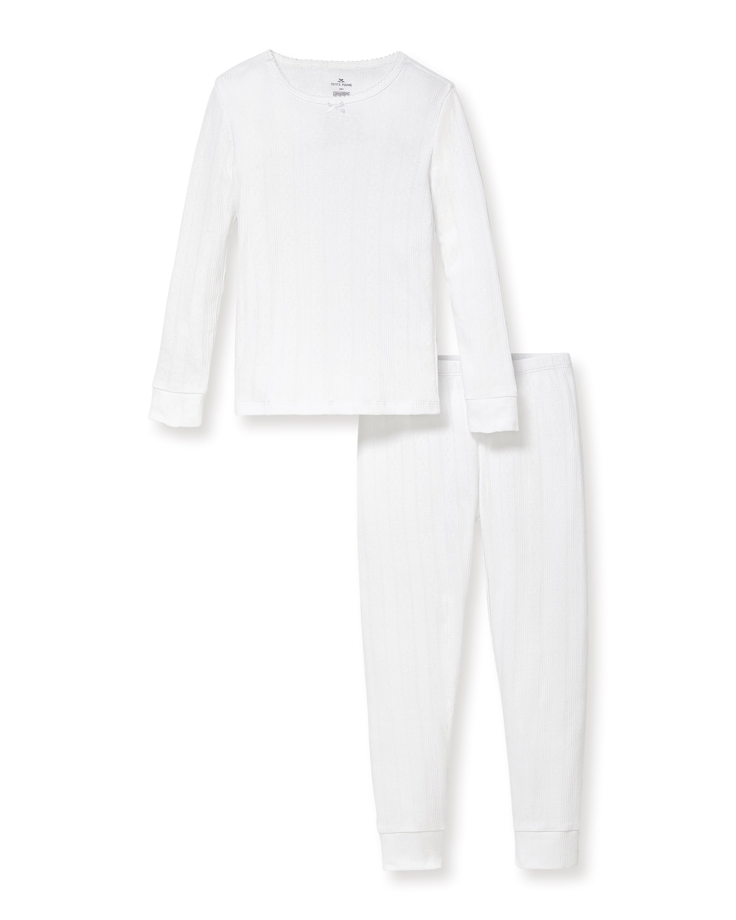 A happy child with long blonde hair stands barefoot against a plain background, wearing the Kid's Pointelle Snug Fit Pajama Set in White—long-sleeve top and matching pants—with one hand behind their head and the other arm relaxed.