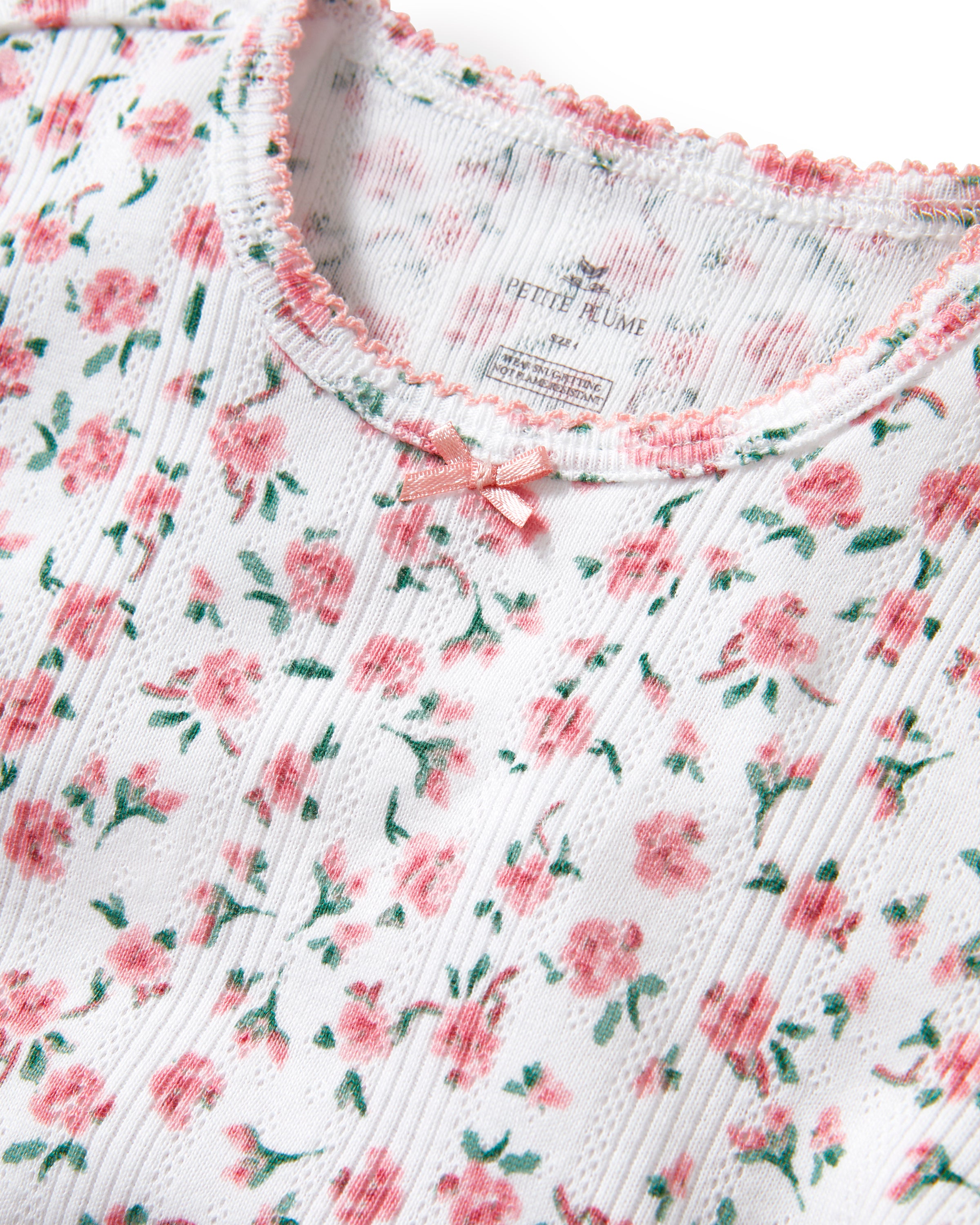A young girl with curly hair stands against a plain background, wearing the Kid's Pointelle Snug Fit Pajama Short Set in Pink Floral, featuring a matching short-sleeve top and shorts decorated with a pink and red floral pattern.