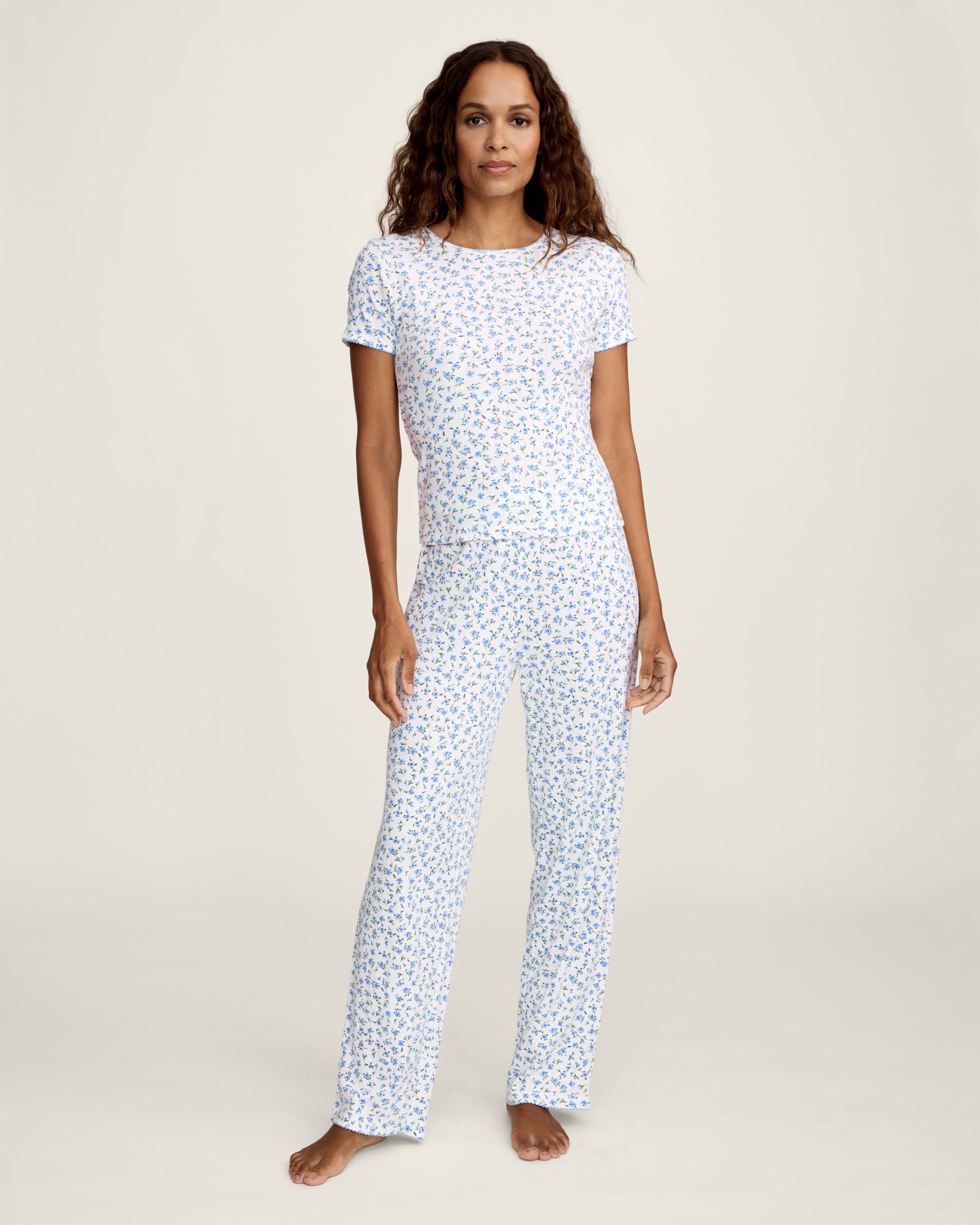 A woman stands barefoot against a plain background, wearing the Women's Pointelle Pant in Blue Floral paired with a matching short-sleeve pajama top featuring a white base and small blue floral print.