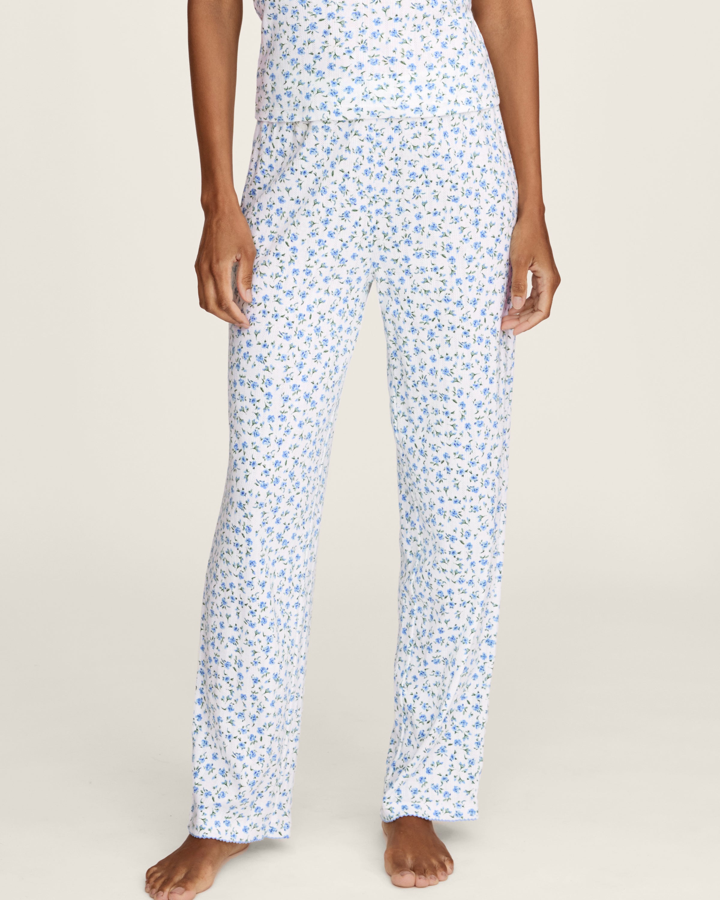 A woman stands barefoot against a plain background, wearing the Women's Pointelle Pant in Blue Floral paired with a matching short-sleeve pajama top featuring a white base and small blue floral print.