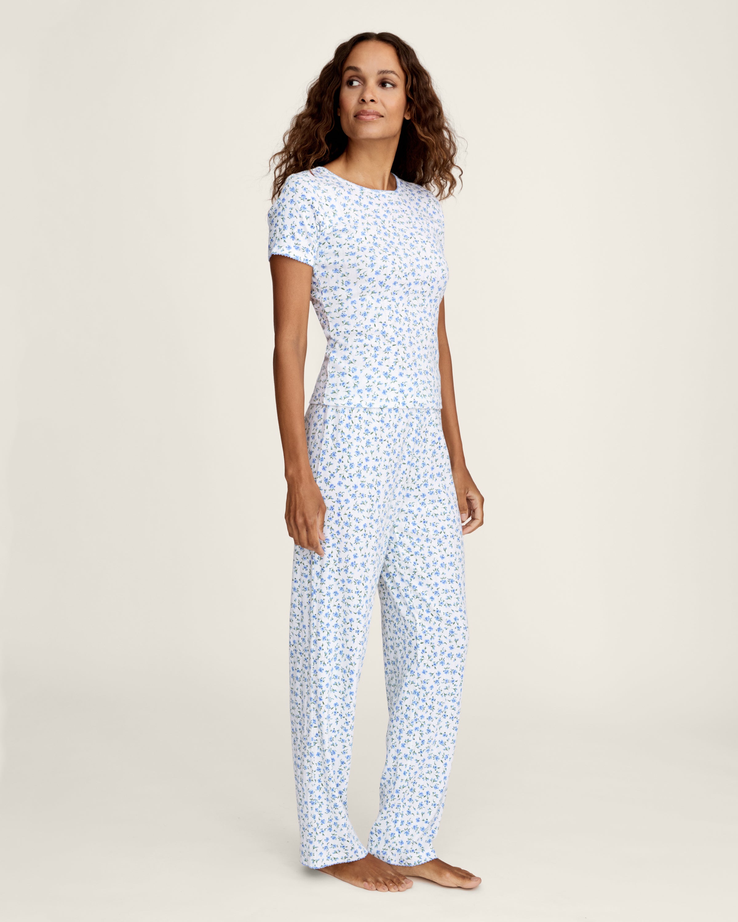A woman stands barefoot against a plain background, wearing the Women's Pointelle Pant in Blue Floral paired with a matching short-sleeve pajama top featuring a white base and small blue floral print.