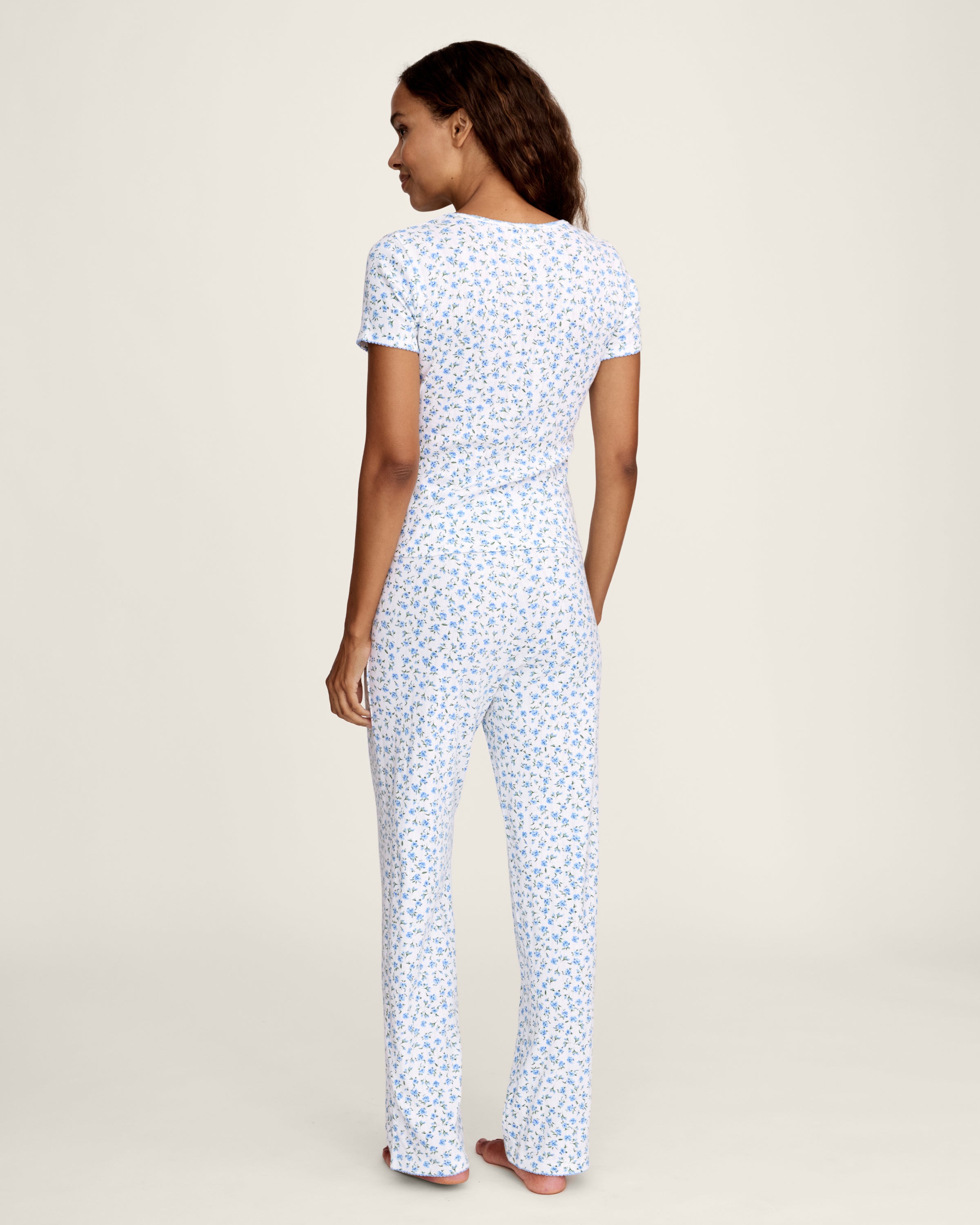 A woman stands barefoot against a plain background, wearing the Women's Pointelle Pant in Blue Floral paired with a matching short-sleeve pajama top featuring a white base and small blue floral print.