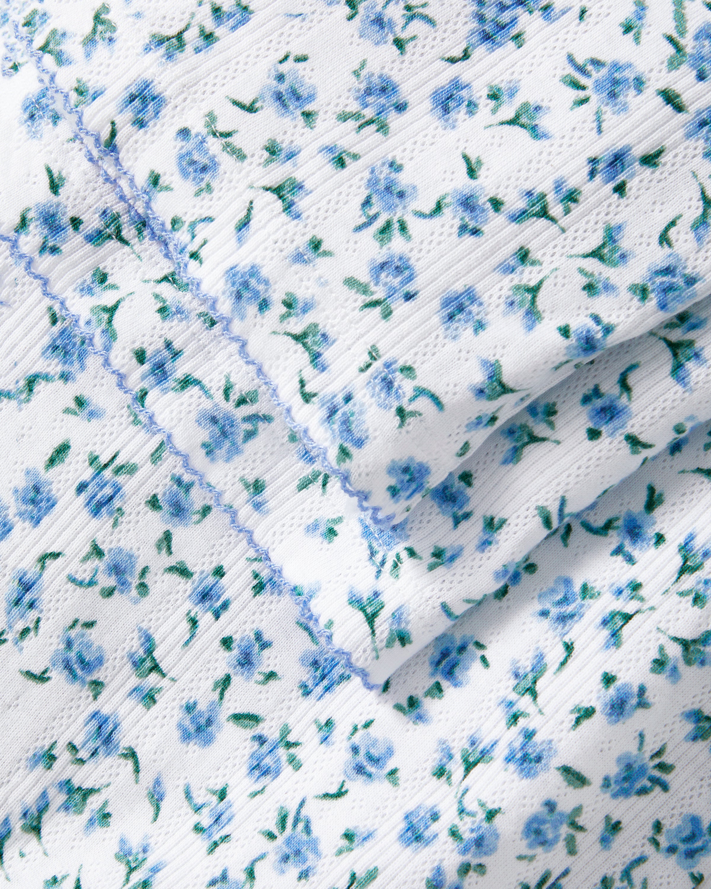 A woman stands barefoot against a plain background, wearing the Women's Pointelle Pant in Blue Floral paired with a matching short-sleeve pajama top featuring a white base and small blue floral print.