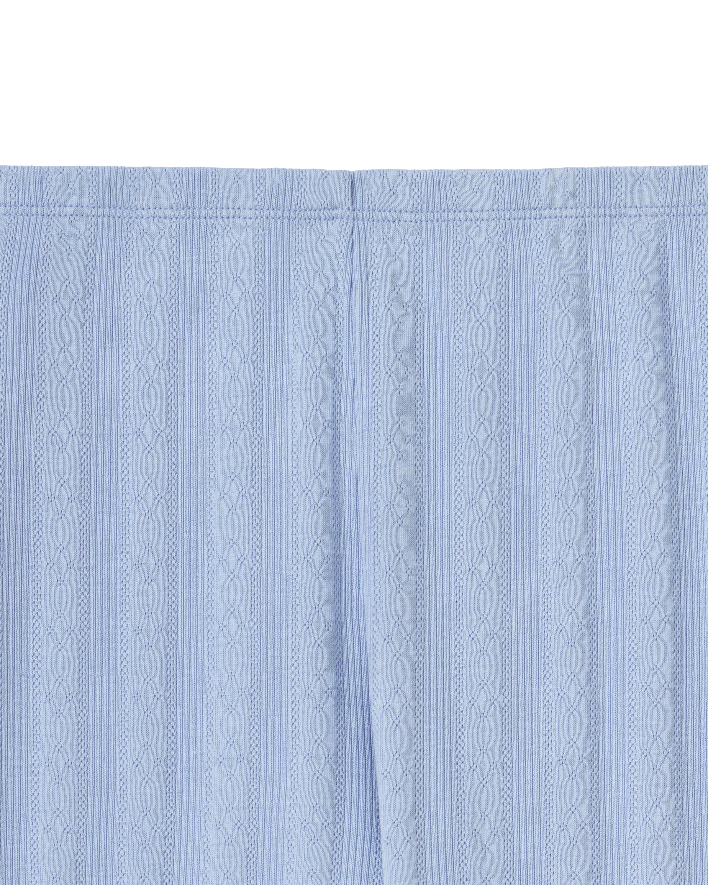 A woman stands barefoot in Women's Pointelle Pant in Periwinkle, paired with a matching short-sleeve top. She has wavy brown hair and gazes to the side against a plain, light background.