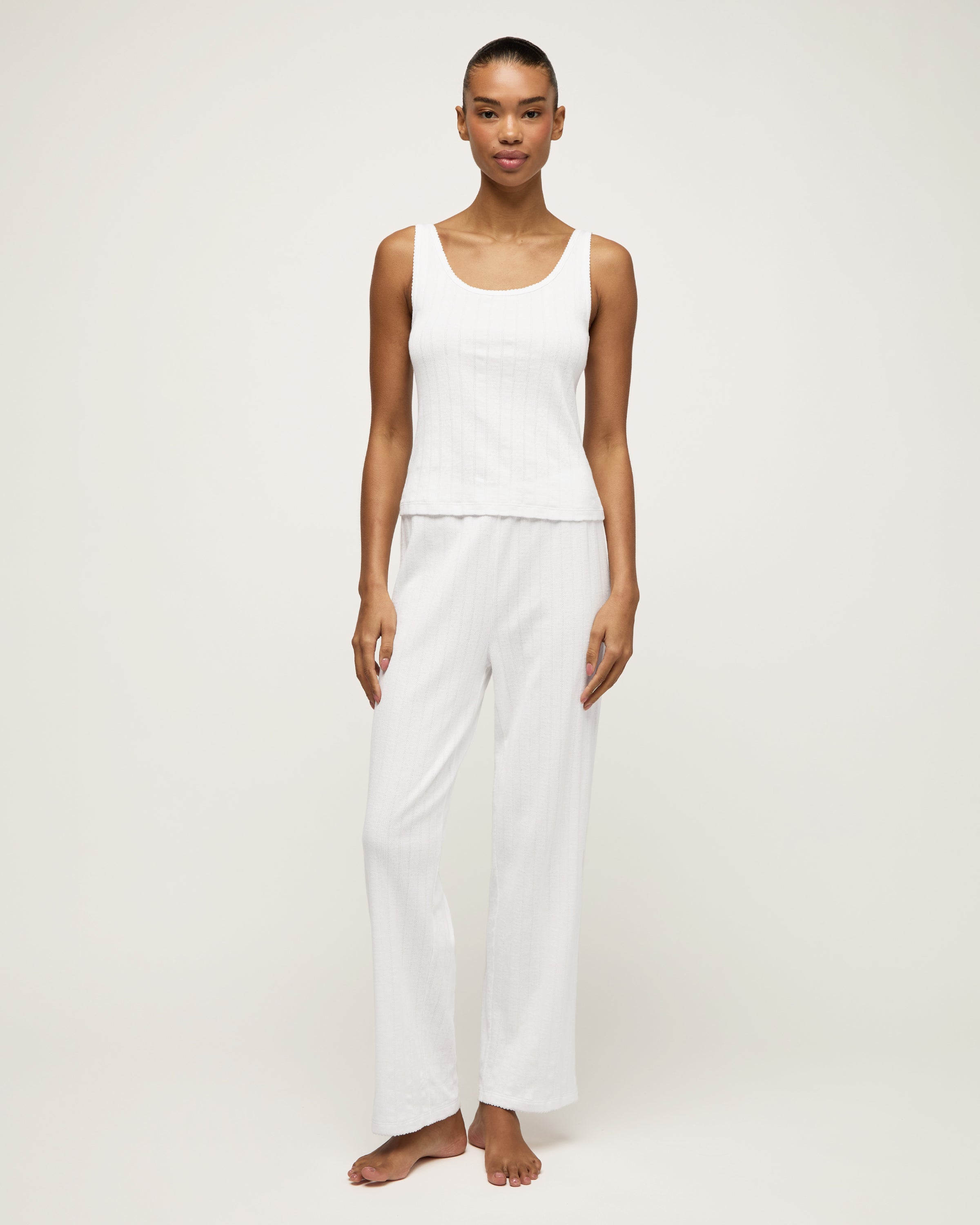 A woman stands barefoot on a light background wearing the Women's Pointelle Pant in White, paired with a sleeveless white tank top, posing with a relaxed and neutral expression.