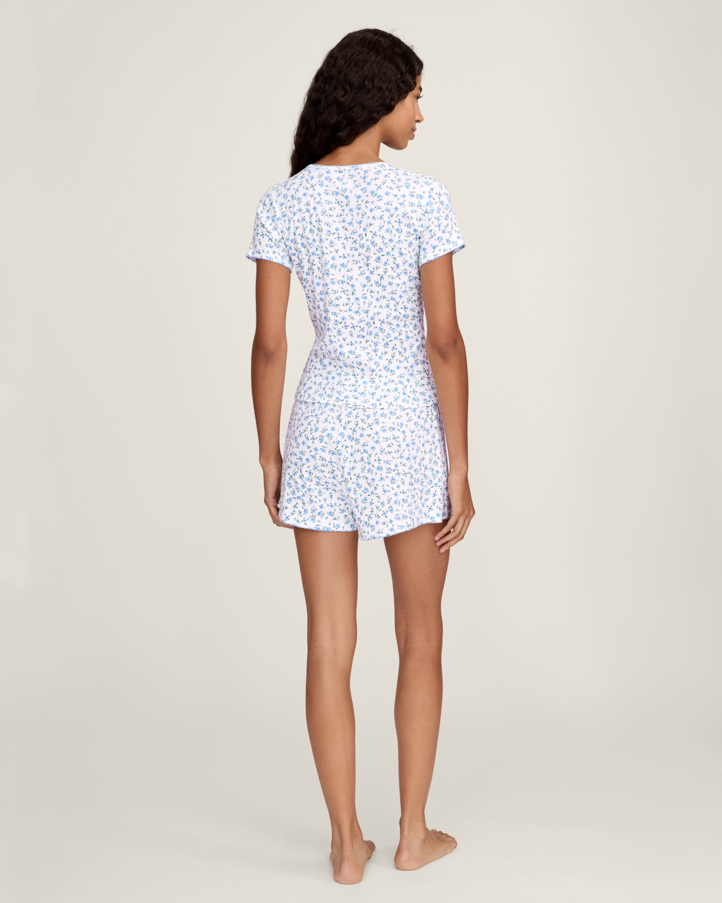 A person stands barefoot against a plain background, wearing Women's Pointelle Shorts in Blue Floral with a matching short-sleeve top. They have long, wavy hair and a relaxed, neutral expression.