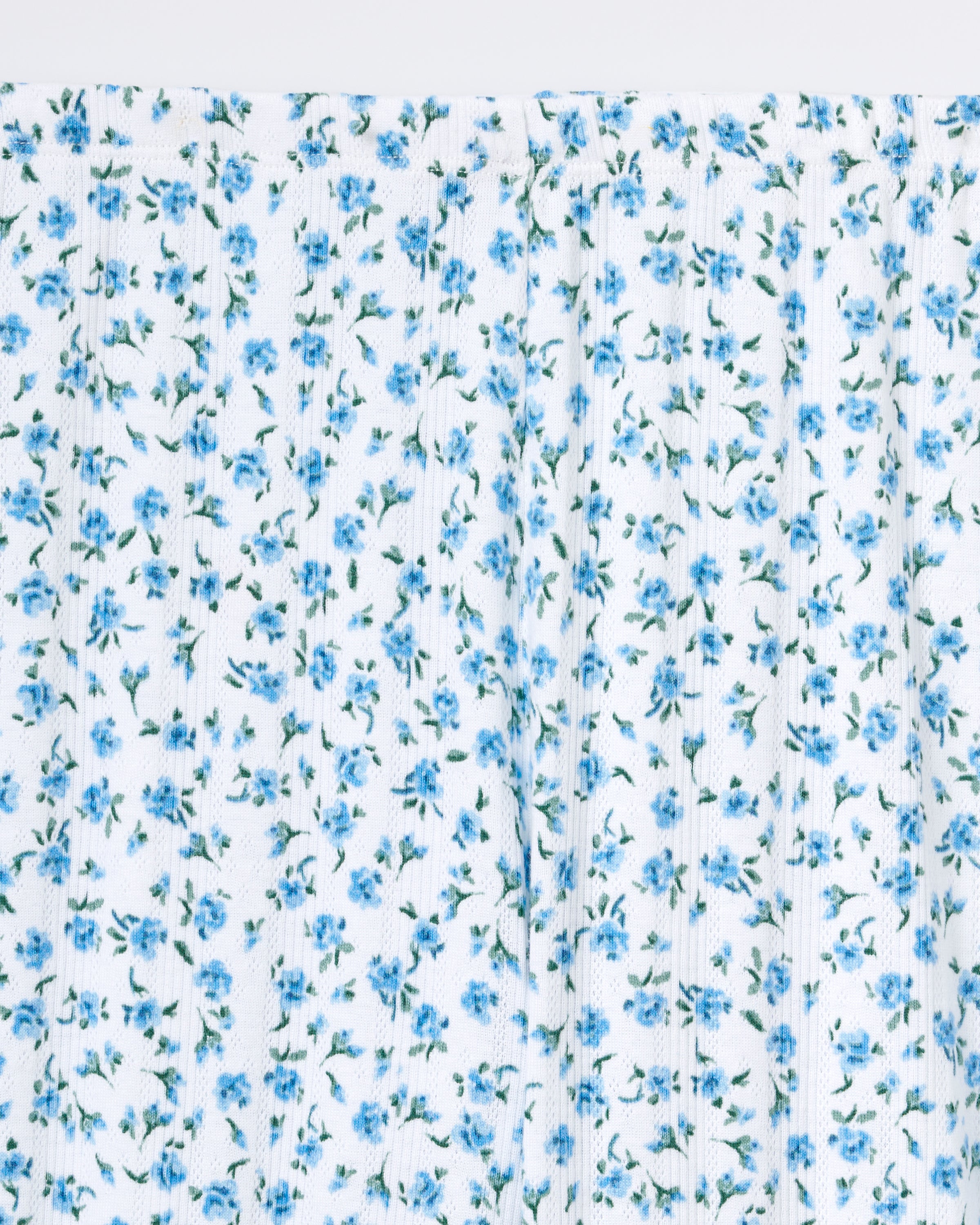 A person stands barefoot against a plain background, wearing Women's Pointelle Shorts in Blue Floral with a matching short-sleeve top. They have long, wavy hair and a relaxed, neutral expression.