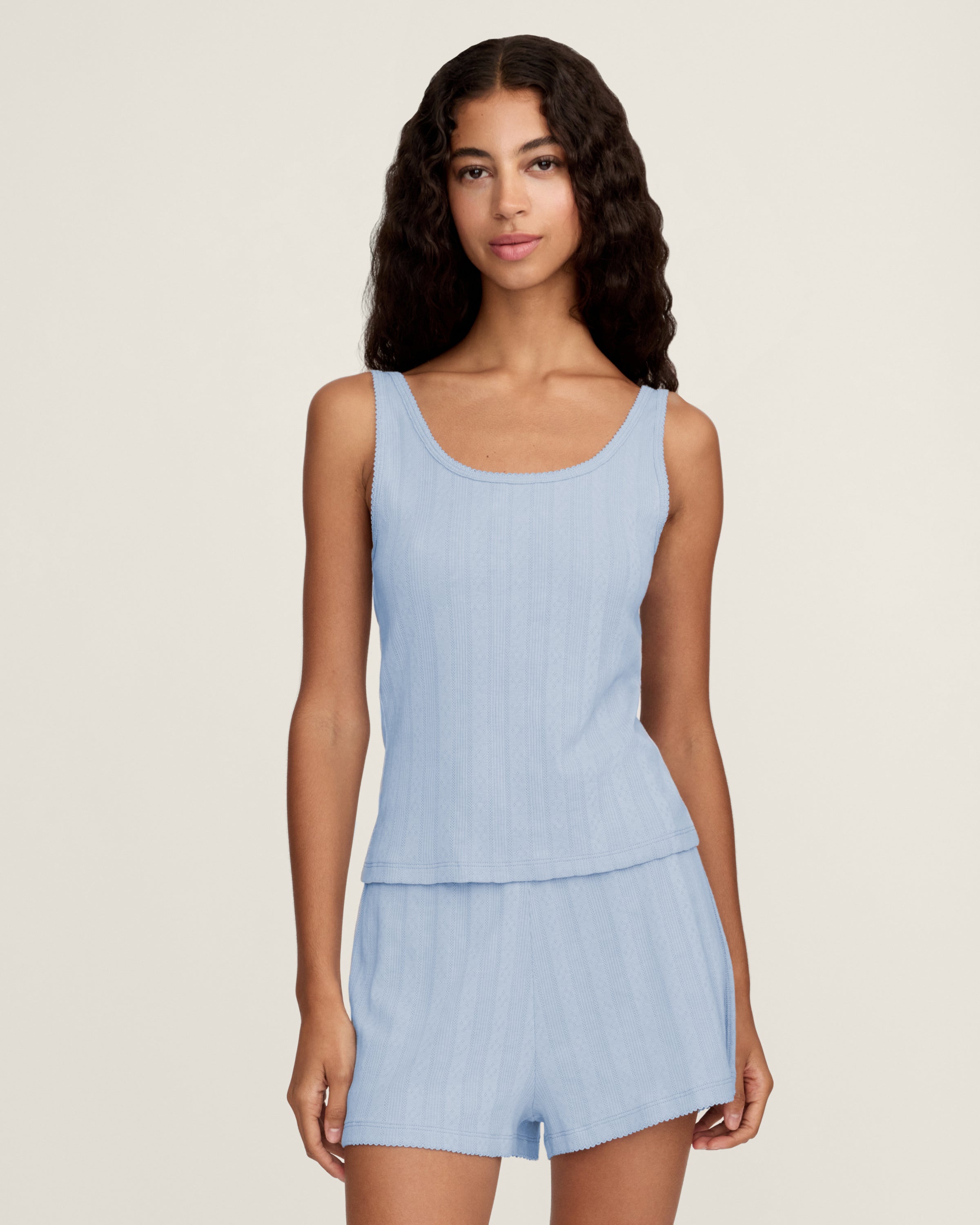 A woman with long wavy hair stands barefoot on a plain background, wearing Women's Pointelle Shorts in Periwinkle and a matching light blue ribbed tank top. She has a relaxed, neutral expression.
