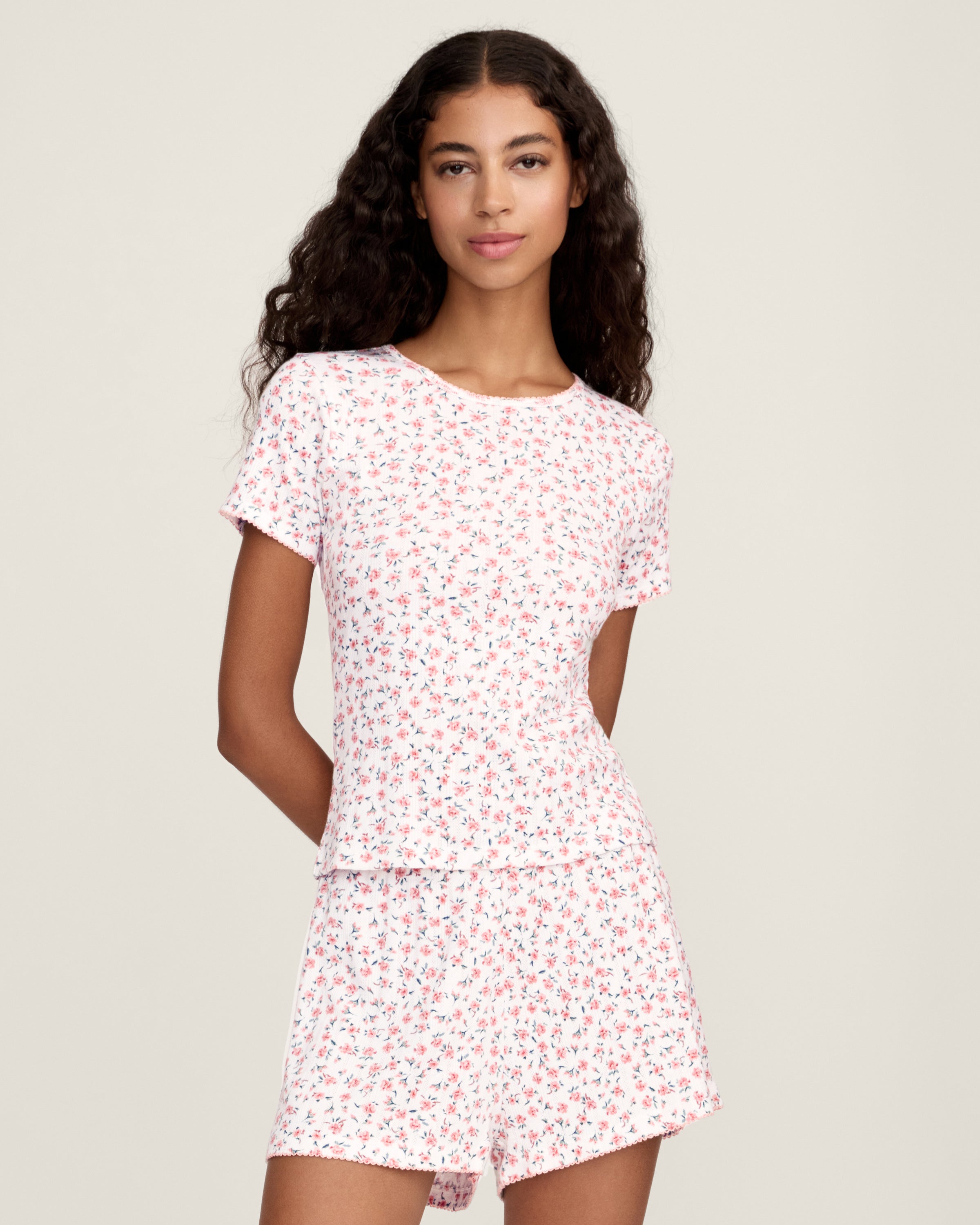 A woman with long curly hair and a relaxed expression stands barefoot against a neutral background, wearing Women's Pointelle Shorts in Pink Floral as part of a matching short-sleeved pajama set.
