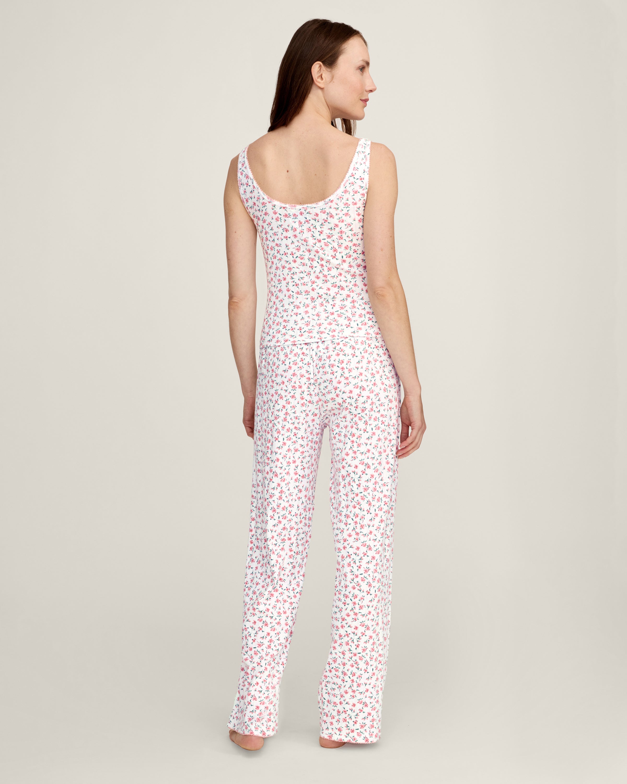 A woman with long brown hair smiles softly, wearing the Women's Pointelle Tank in Pink Floral with matching pants, standing against a plain light background.