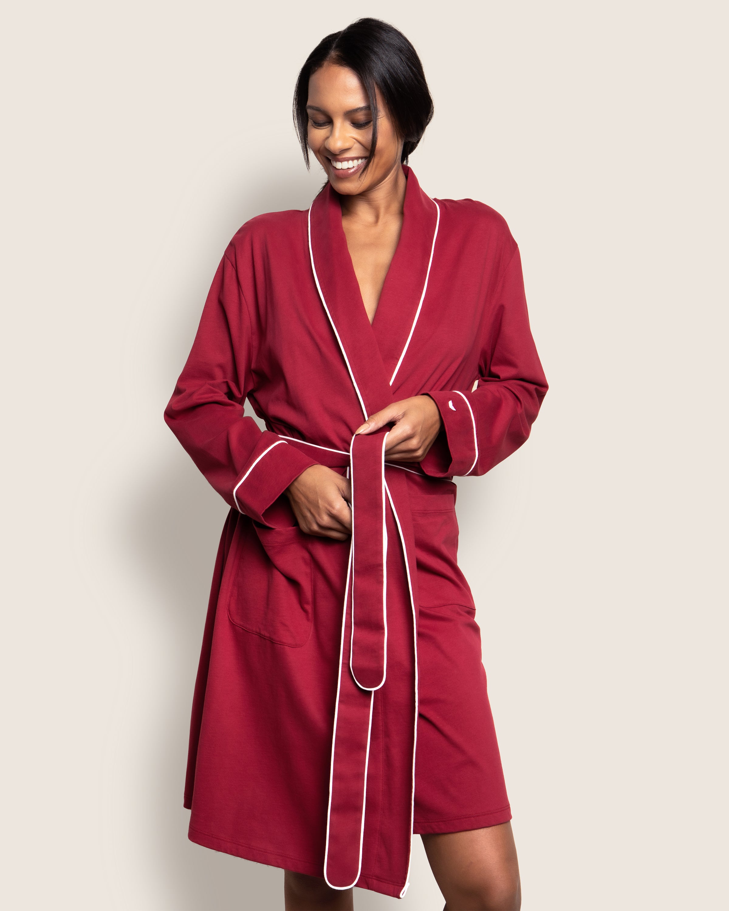 A person smiling, wearing Petite Plumes Womens Pima Robe in Bordeaux with white trim, ties the belt around their waist. The background is plain and light-colored.