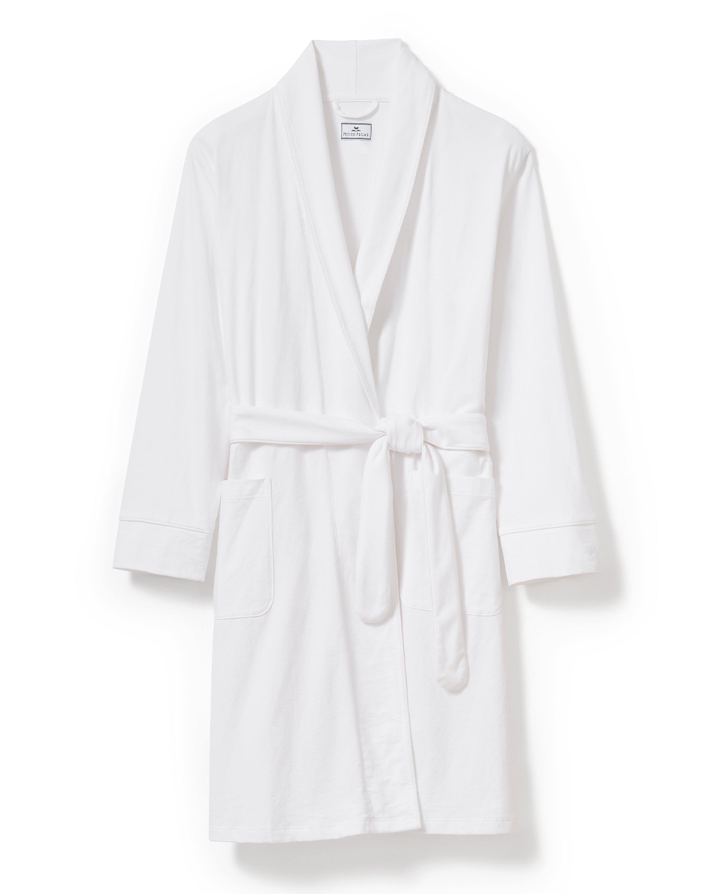 A person with curly hair is wearing a Petite Plume Womens Pima Robe in White, crafted from luxurious Peruvian Pima cotton. With hands in pockets, they stand against a plain light background, looking forward with a calm expression.