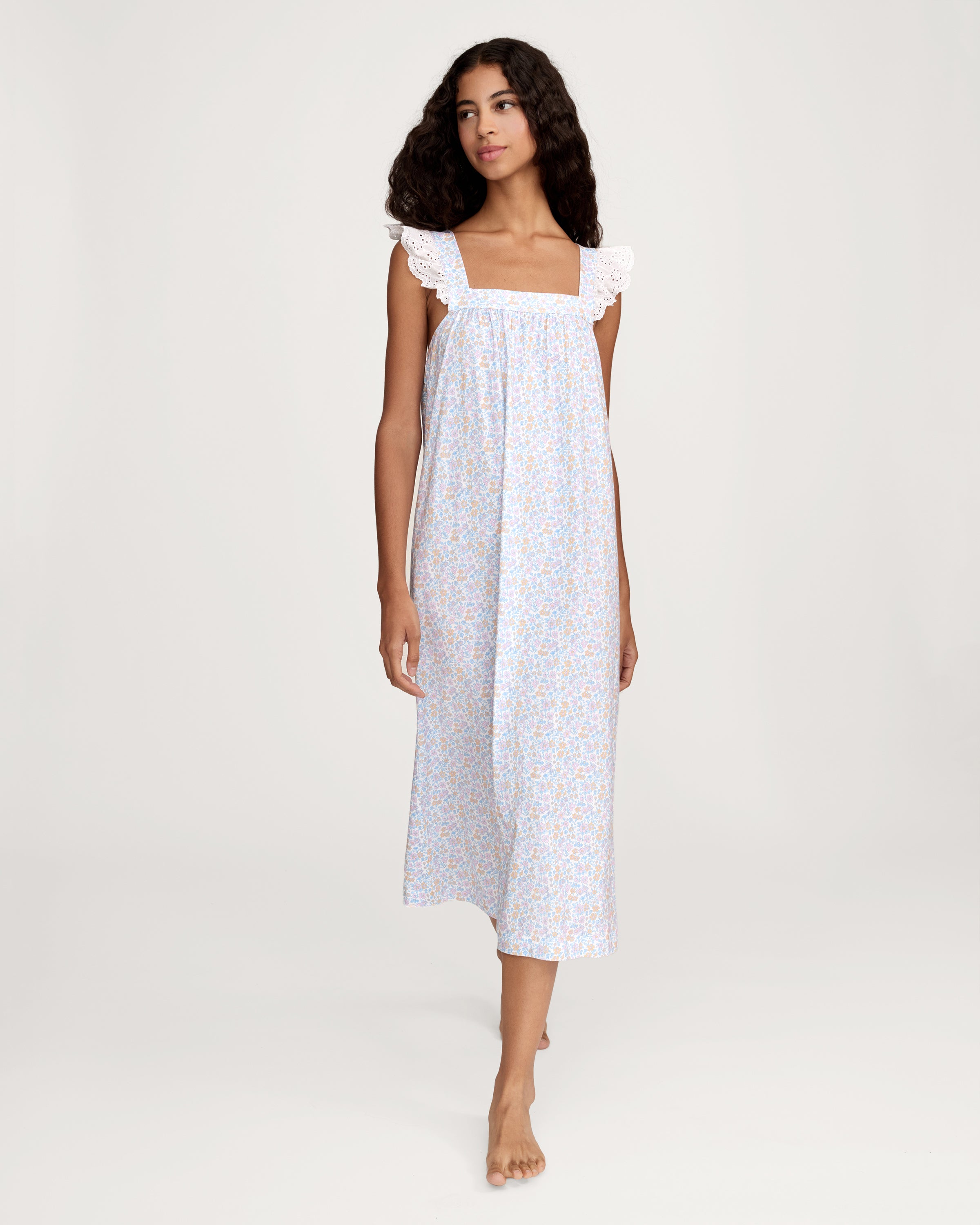 A woman with long curly hair stands barefoot on a plain light background, wearing the Women's Lightweight Pima Clara Nightgown in Fleur Blanche—an ankle-length, sleeveless gown with dainty floral print and lace-trimmed straps.