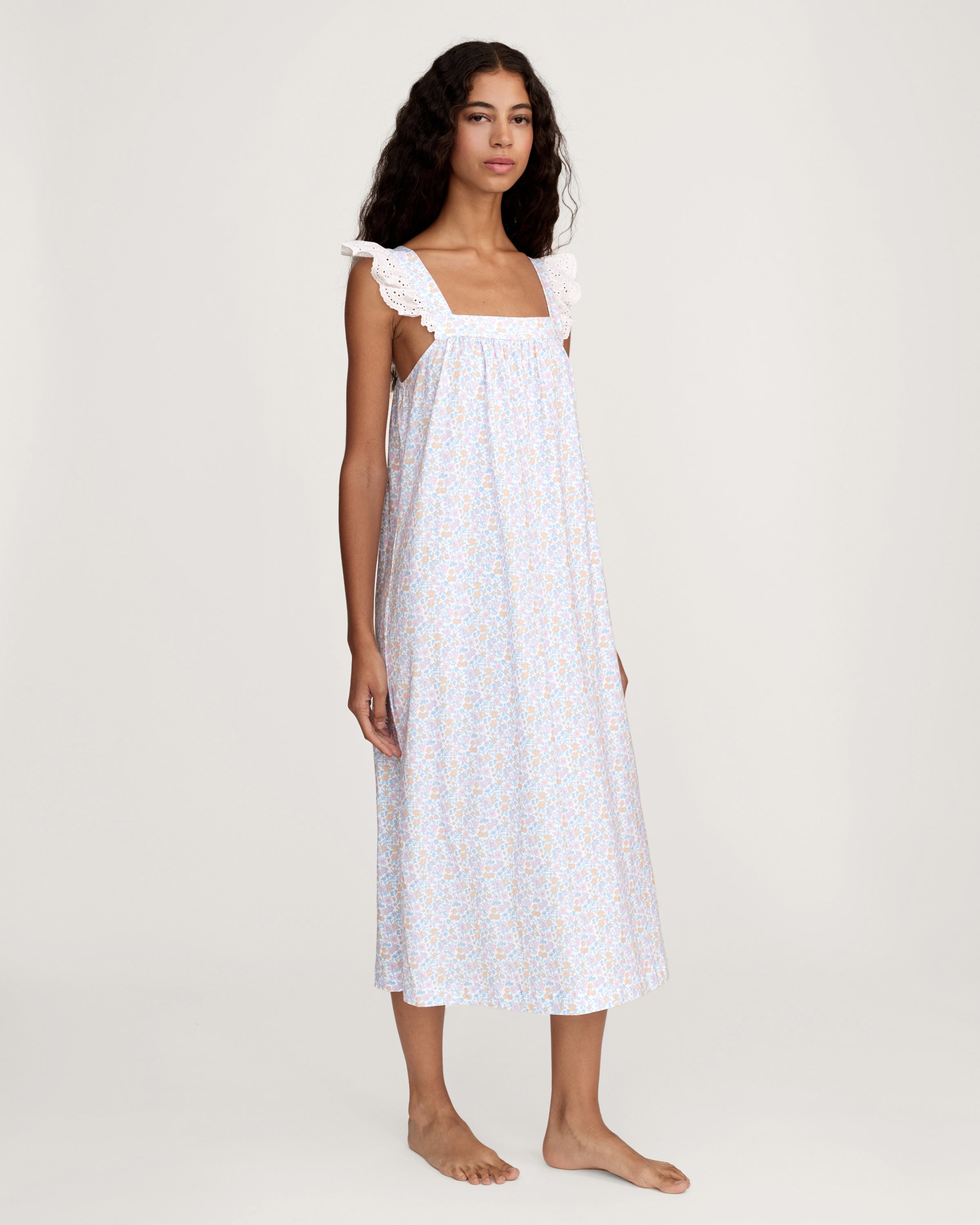 A woman with long curly hair stands barefoot on a plain light background, wearing the Women's Lightweight Pima Clara Nightgown in Fleur Blanche—an ankle-length, sleeveless gown with dainty floral print and lace-trimmed straps.
