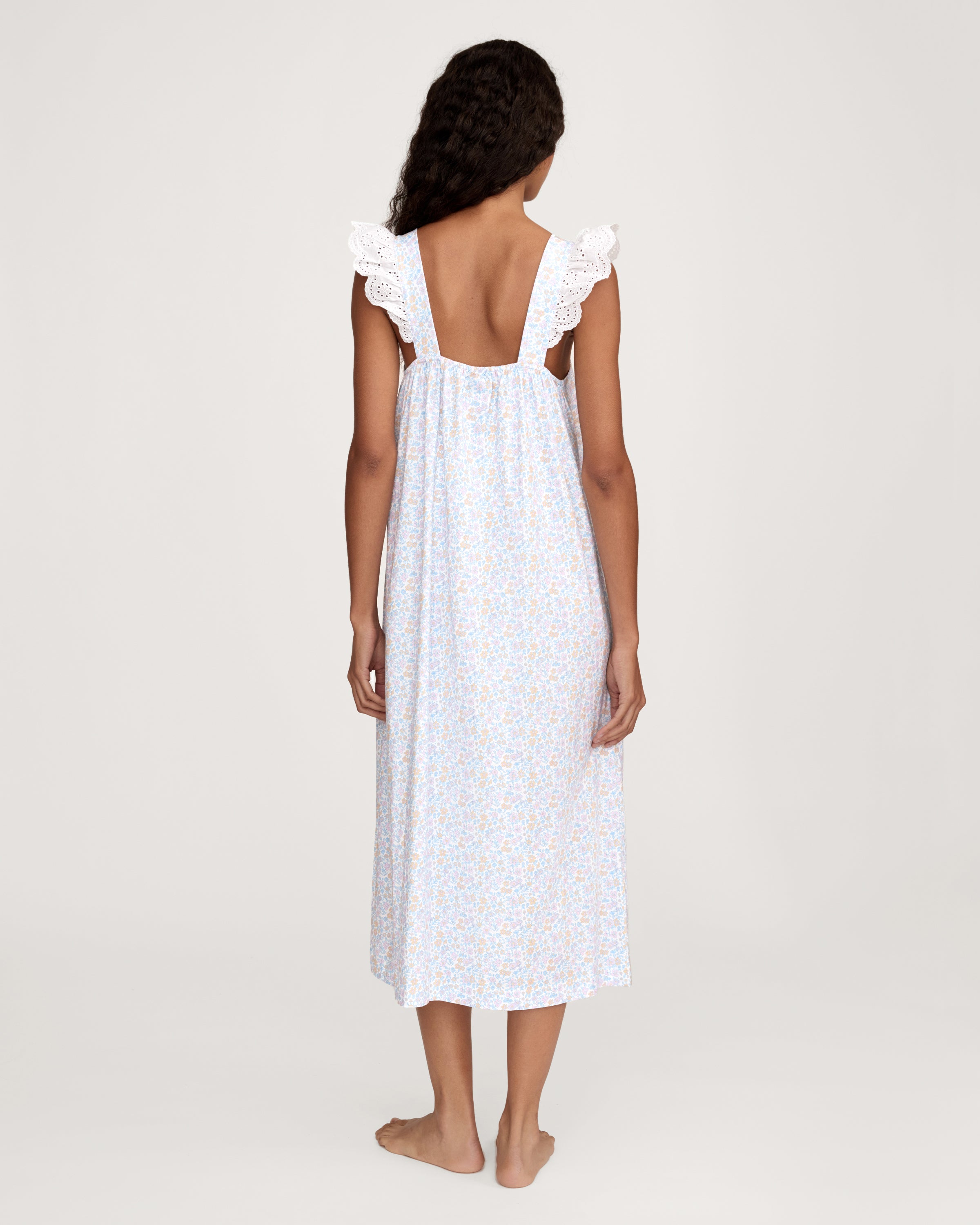 A woman with long curly hair stands barefoot on a plain light background, wearing the Women's Lightweight Pima Clara Nightgown in Fleur Blanche—an ankle-length, sleeveless gown with dainty floral print and lace-trimmed straps.