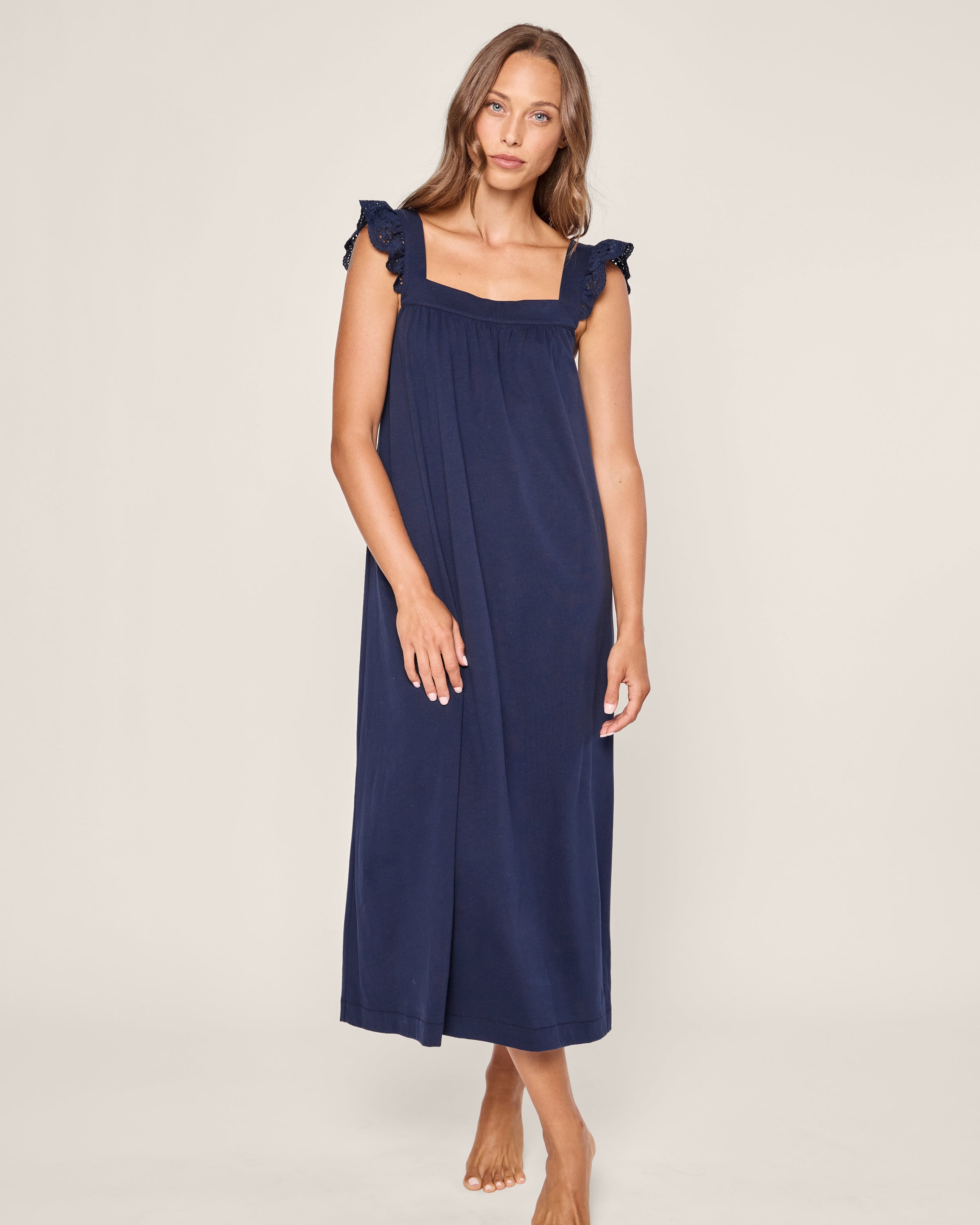 A woman wearing the Petite Plume Womens Pima Clara Nightgown in navy, showcasing its Peruvian Pima cotton fabric, stands barefoot against a neutral background. She has long brown hair and a relaxed posture, with her left hand gently resting on her hip.